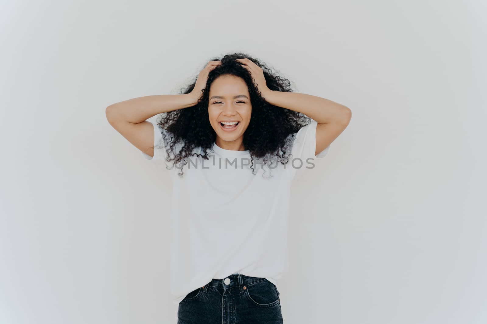 Photo of overjoyed curly haired woman laughs happily, has fun, dressed in white t shirt and jeans, smiles broadly, isolated over white background. People, emotions, happiness, ethnicity concept by vkstock