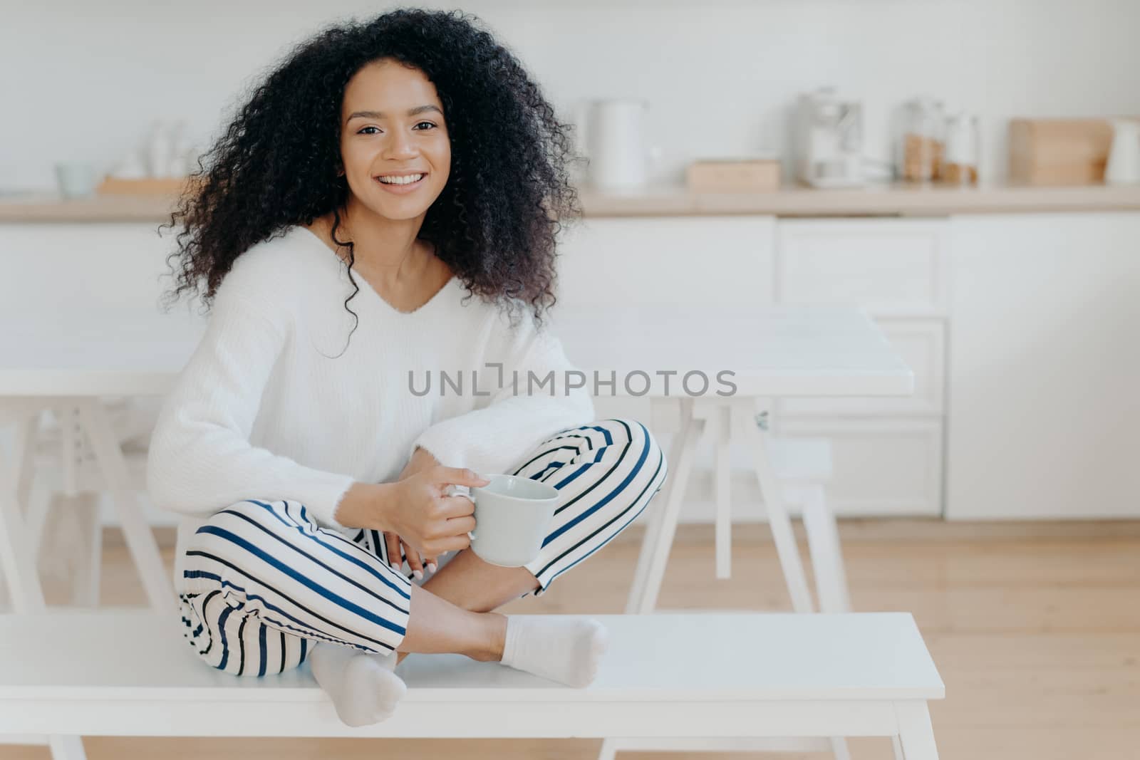 Smiling relaxed African American female sits crossed legs on bench against kitchen interior, wears white sweater and striped pants, drinks hot beverage, enjoys domestic atmosphere. Coffee time