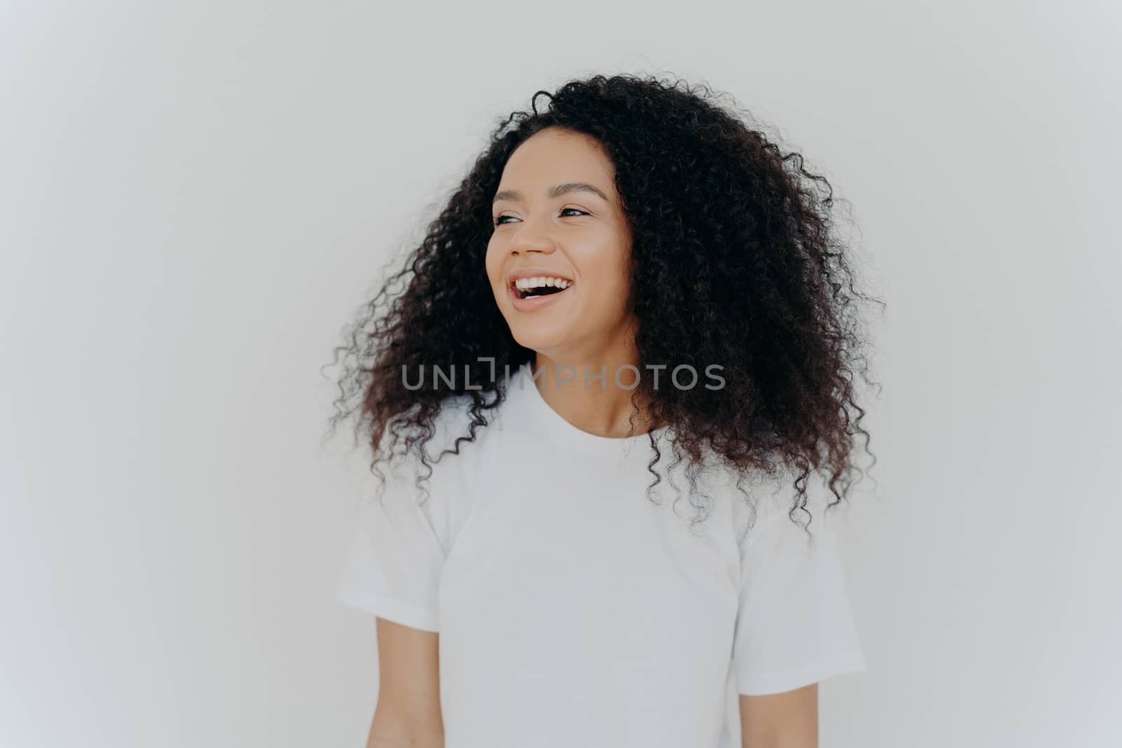 Portrait of beautiful curly adult woman turns gaze aside, has glad expression, smiles and laughs at something funny, dressed in white t shirt, models indoor, feels free, energized and relieved, by vkstock