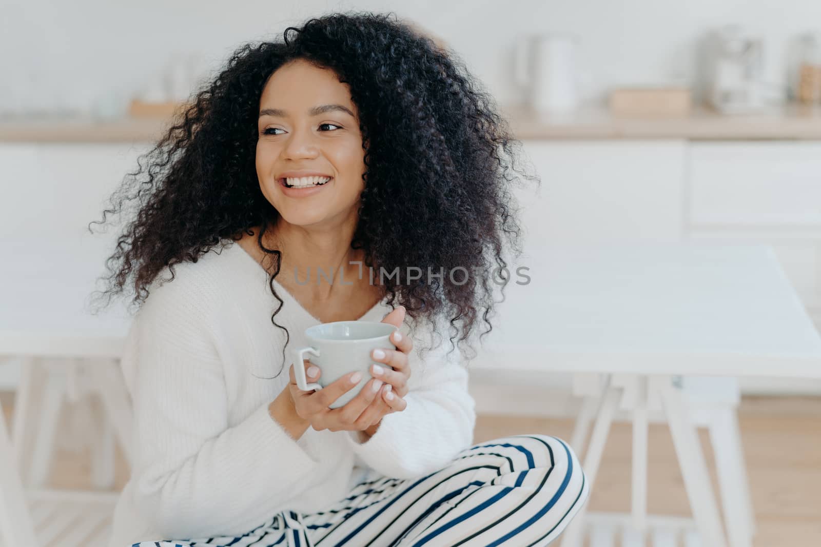 Photo of cute cheerful woman with bushy curly hair looks away with smile, holds mug of coffee, wears casual clothing, enjoys cozy calm morning, savors taste. Afro lay in kitchen during day off by vkstock