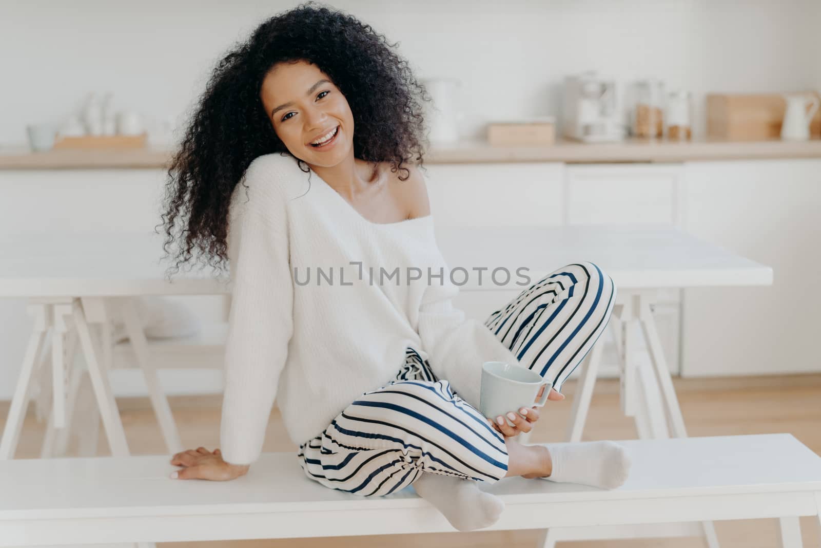Pleased young girlfriend with Afro curly hairstyle, sits crossed legs on bench, enjoys morning coffee and domestic atmosphere, poses against kitchen background, spends time alone, has breakfast