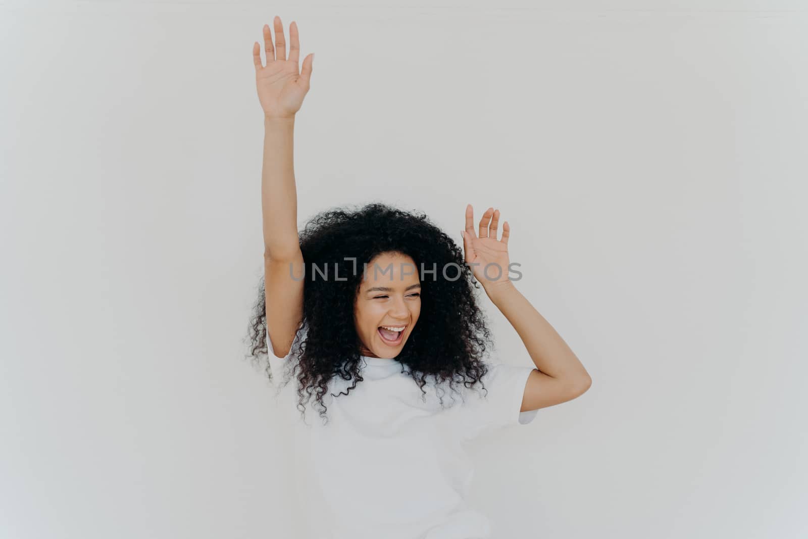 Joyful curly African American woman raises hands up, laughs from happiness, celebrates triumph, rejoices success, wears white t shirt, poses indoor, gestures actively, gets unexpected surprise by vkstock