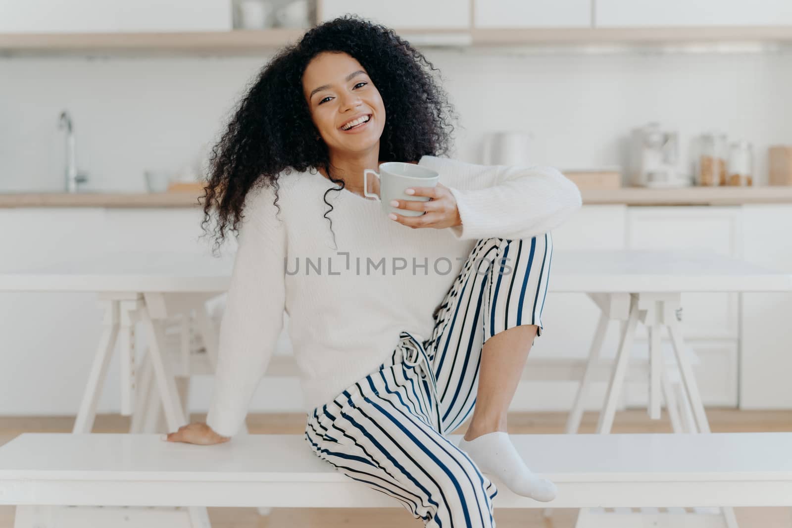 Beautiful young female with Afro haircut, poses on white bench indoor, dressed in stylish clothing, enjoys aromatic beverage, enjoys breakfast at home, poses against blurred kitchen interior by vkstock