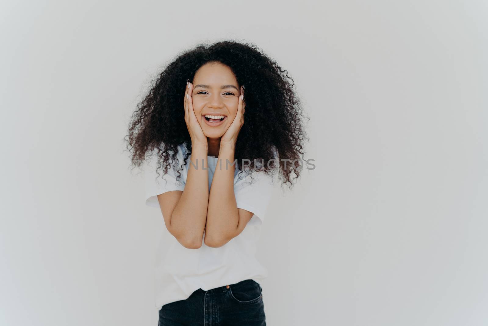 Candid shot of good looking lady with curly hairstyle, smiles cute, keeps both hands on cheeks, shows positive emotions, wears white t shirt, black denim trousers, stands indoor, blank copy space