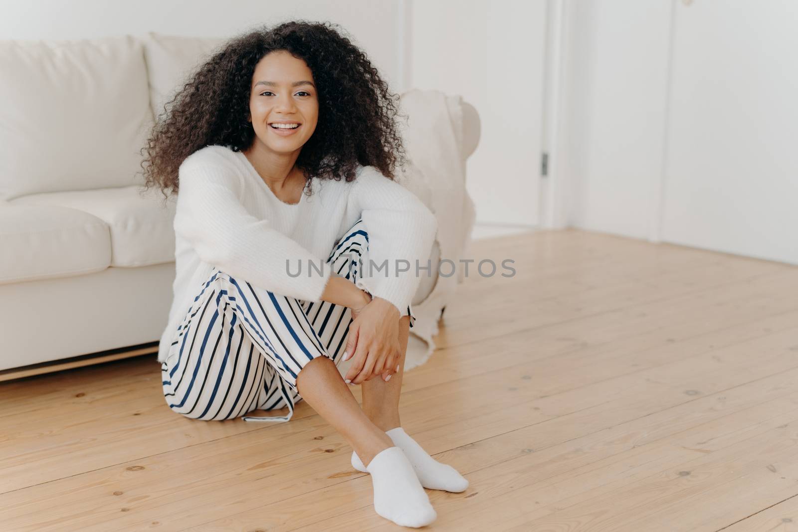 Indoor shot of restful happy African American woman sits on floor in living room, white sofa in background, wears fashionable clothes and socks, expresses positive emorions, rests at home alone by vkstock