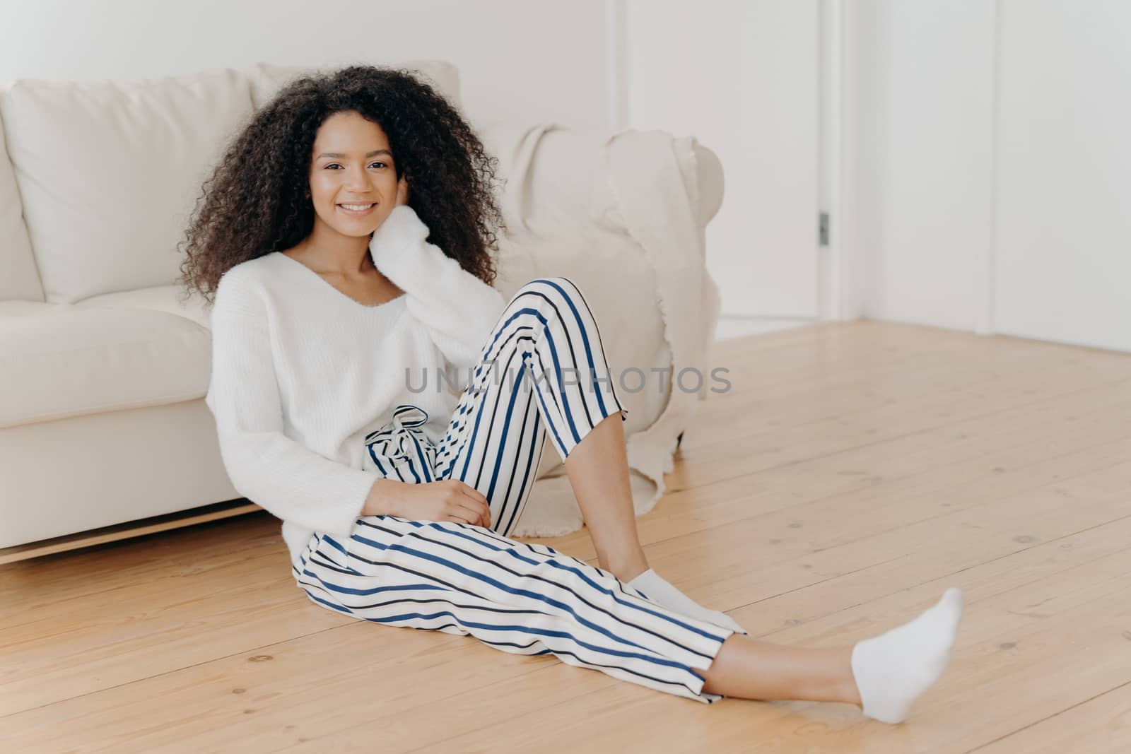 Ethnic girl feels relaxed and satisfied, sits on floor near comfortable sofa in empty room, wears white sweater, striped pants and socks, enjoys domestic atmosphere, enjoys coziness and comfort by vkstock