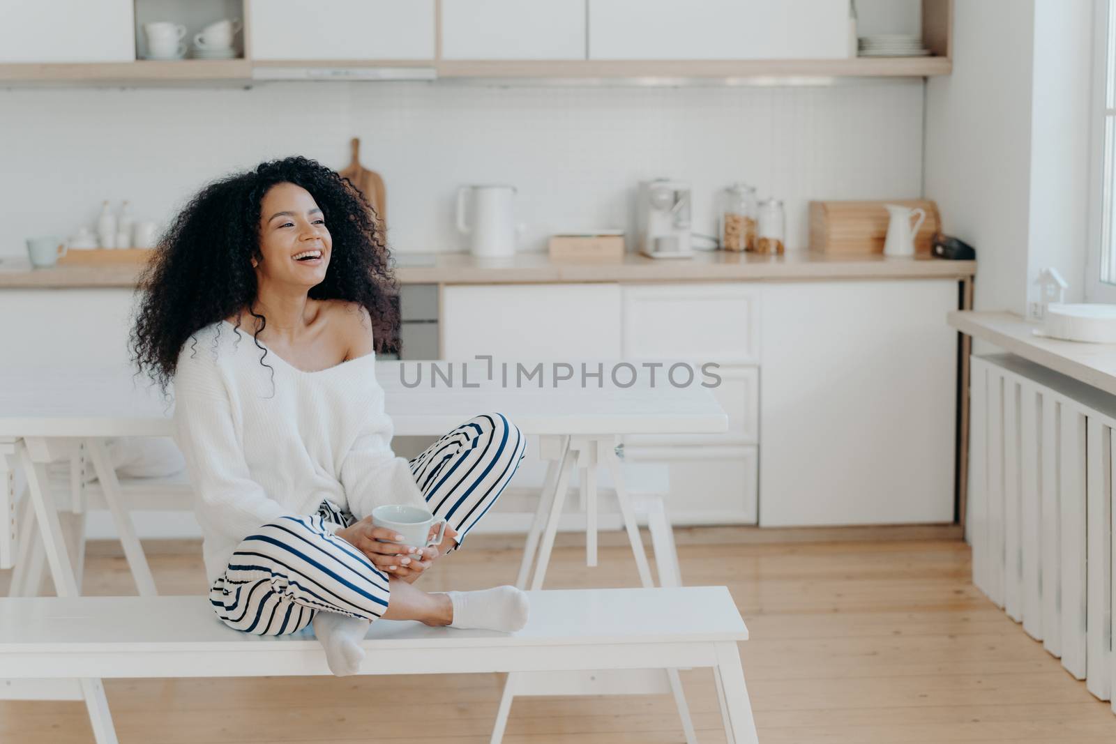 Horizontal shot of overjoyed dark skinned woman laughs pleasantly, drinks coffee, looks out of window in kitchen, dressed in fashionable clothes. Smiling lady with hot tasty beverage, relaxes at home