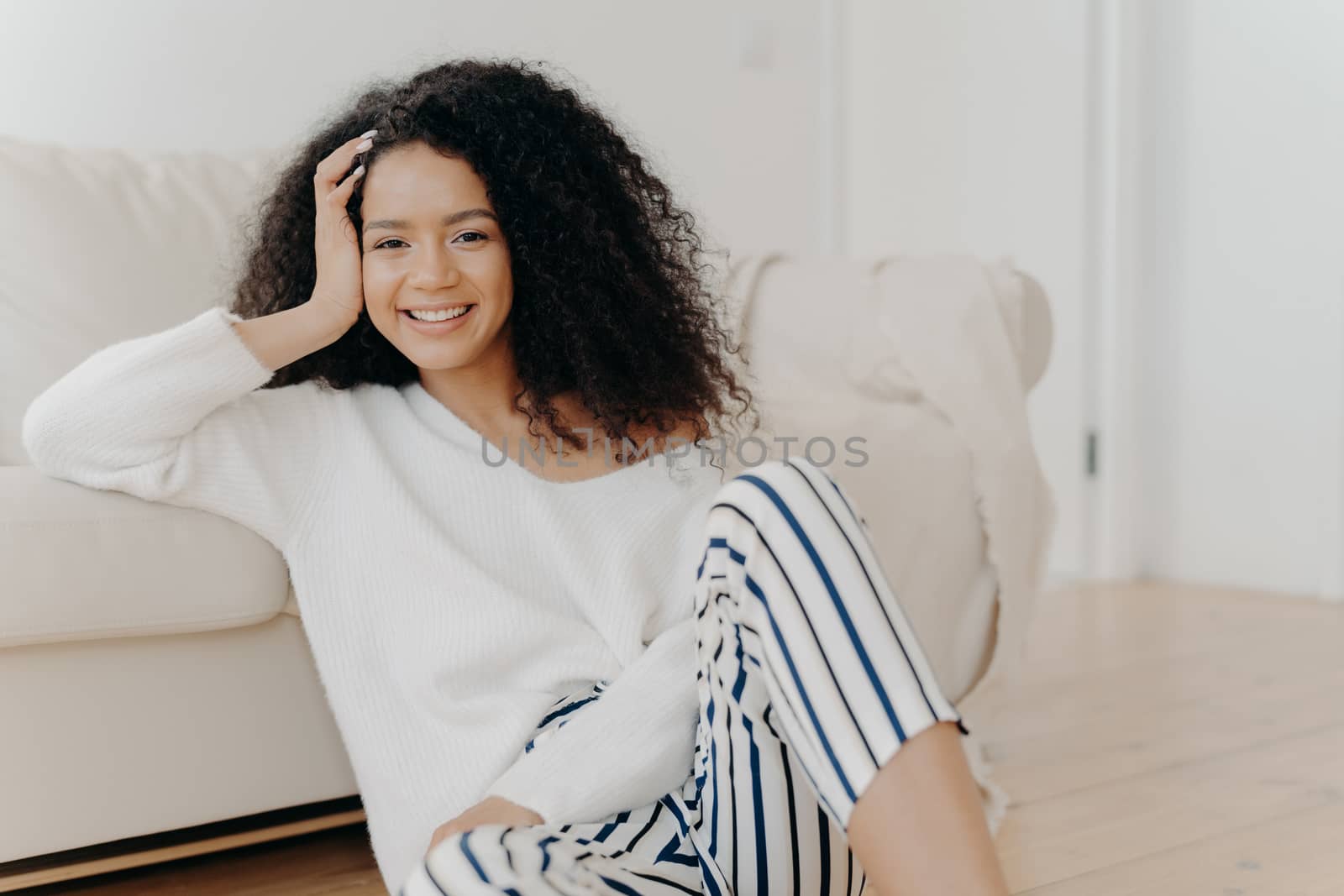 Lovely dark skinned woman with crisp hair, sits on floor near sofa, wears white sweater and striped trousers, enjoys positive emotions, enjoys weekend at home. People, rest, happiness concept by vkstock