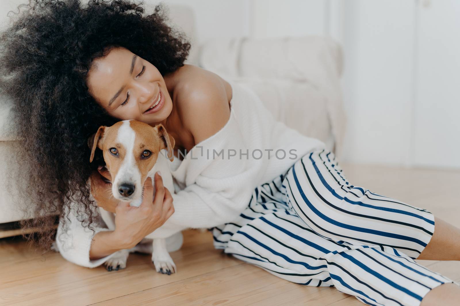 Affectionate young woman hugs dog with love and care, keeps eyes closed from pleasure, smiles gently, has healthy dark skin, poses on floor, petting animal. People, friendship and pets concept by vkstock