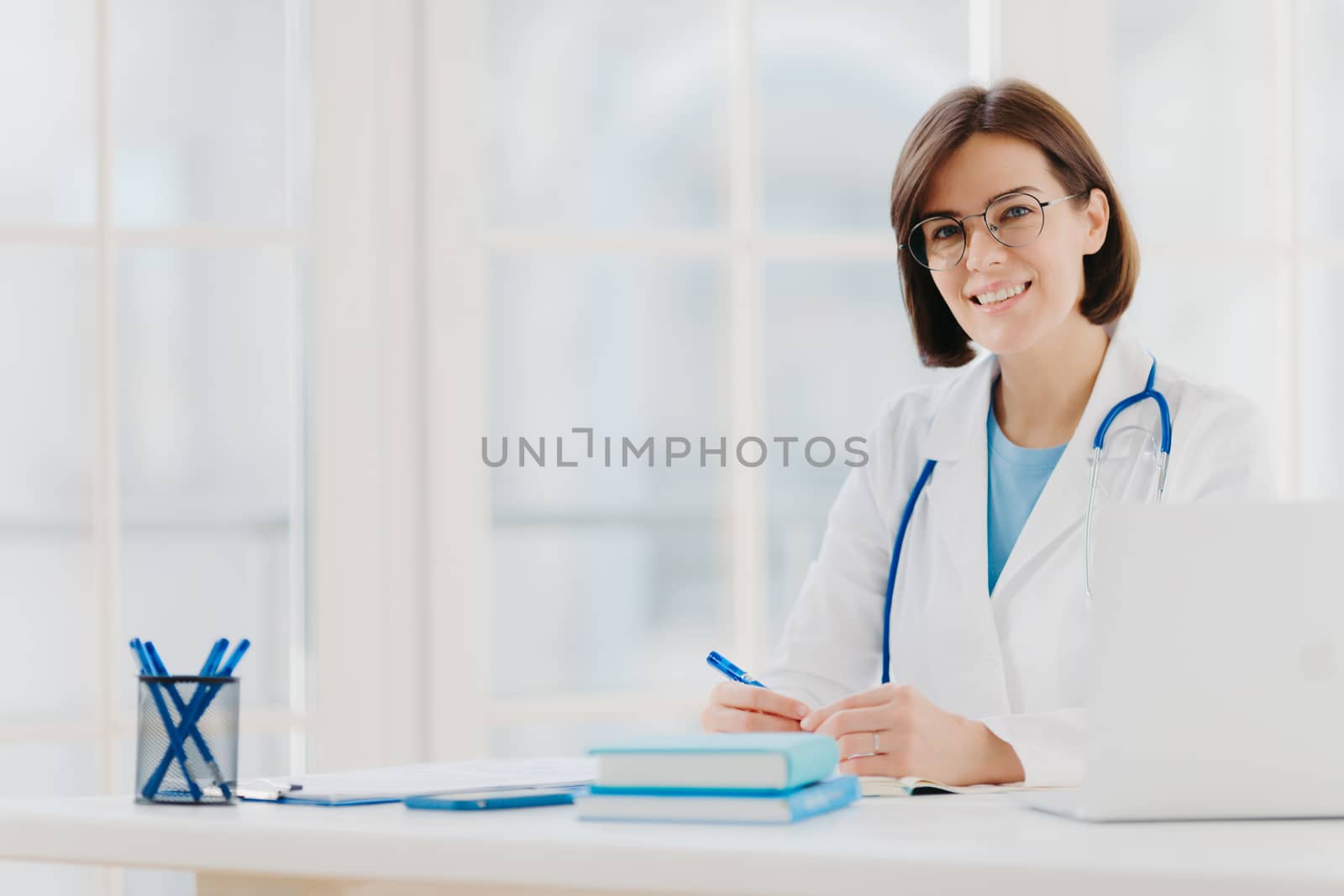 Medical specialist wears white gown with stethoscope, works in hospital, makes notes, poses at own cabinet, sits at table with laptop computer in office smiles happily. Health care and medical concept