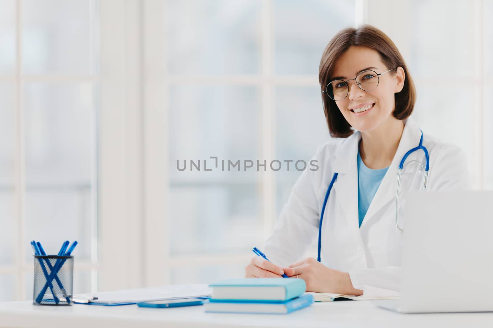 Professional woman doctor writes down notes, poses at desktop in office with laptop, wears white coat, spectacles and phonendoscope around neck, looks through medical documents. Healthcare concept