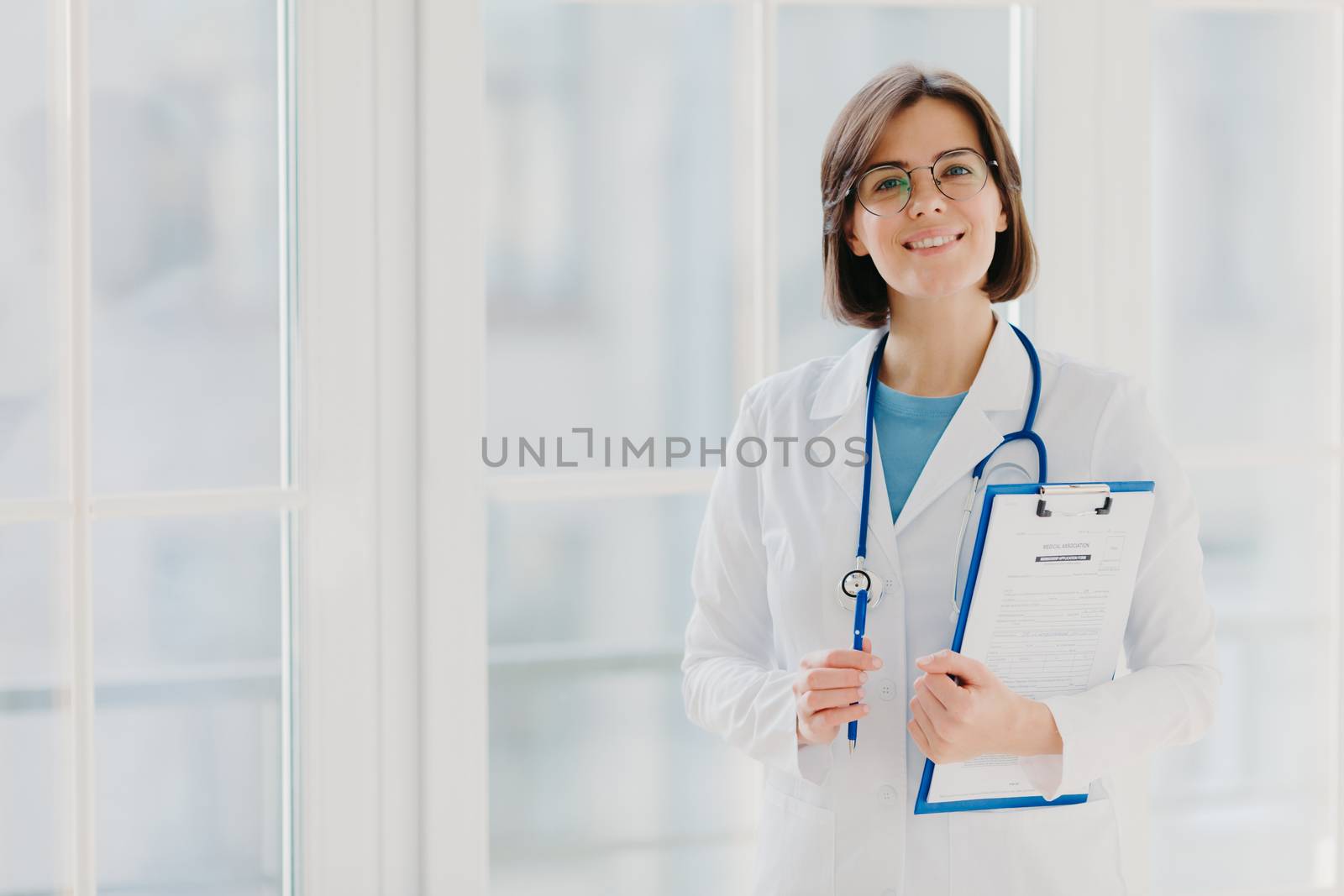 Beautiful smiling intern holds clipboard with written papers and pen, wears white uniform, ready to see patient in own private clinic, stands indoor against white background. Copy space area
