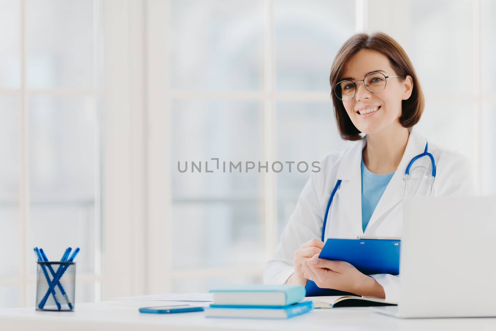 Female doctor writes prescription on special form, works in private clinic, wears white medical gown, ready for seeing patients, poses at workplace. Smiling physician or medical worker holds clipboard by vkstock