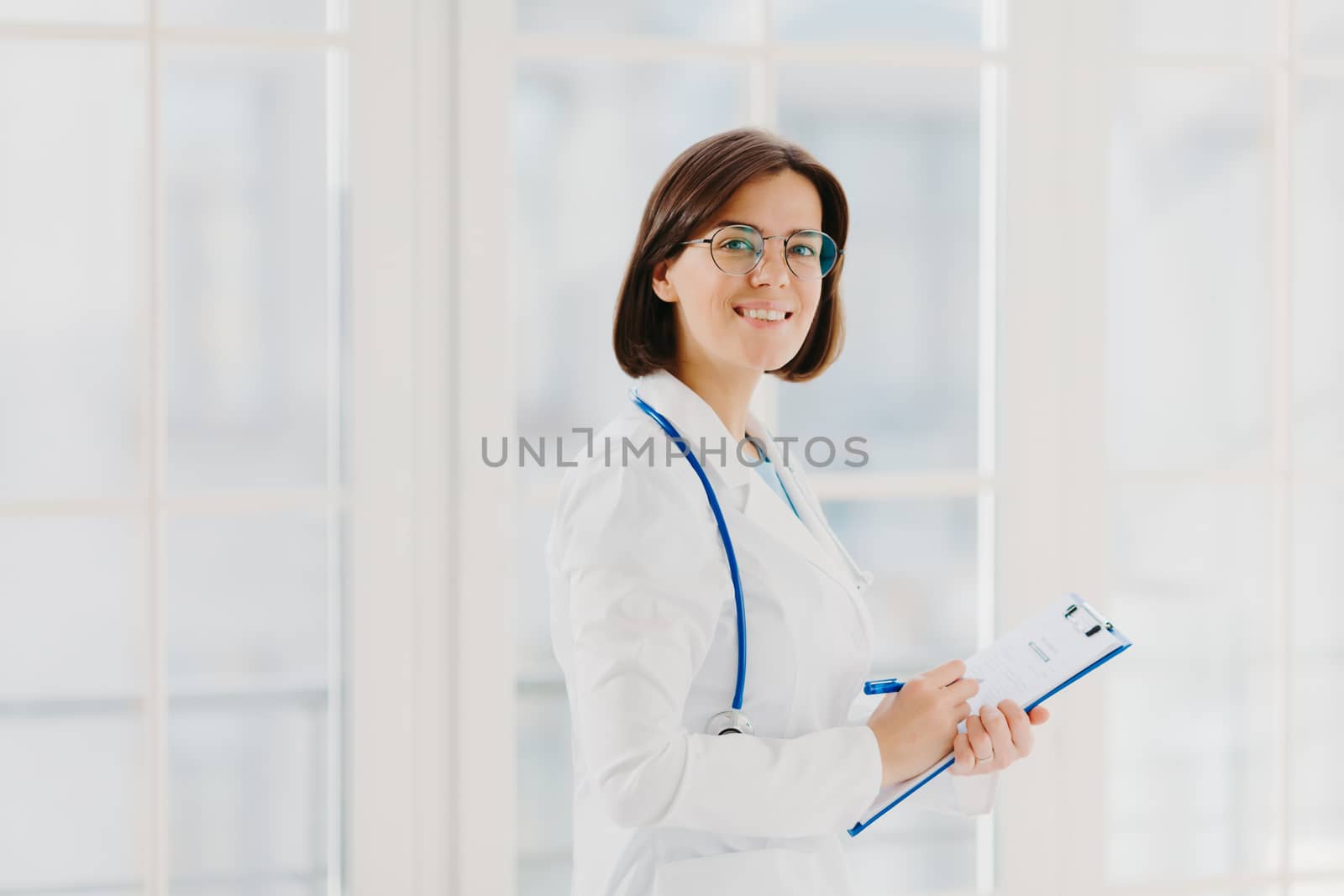 Horizontal shot of female doctor fills up medical form at clipboard, stands indoor, wears round glasses, white gown and stethoscope. General practitioner writes down notes, consults patients