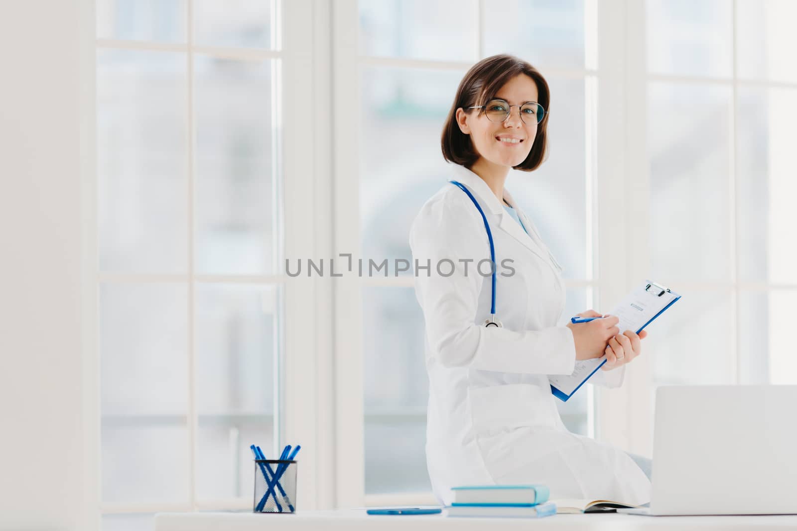 Skilled female physician writes in clipboard, poses at workplace with laptop computer. Professional family doctor ready to give you piece of advice to care about your health, wears medical uniform.