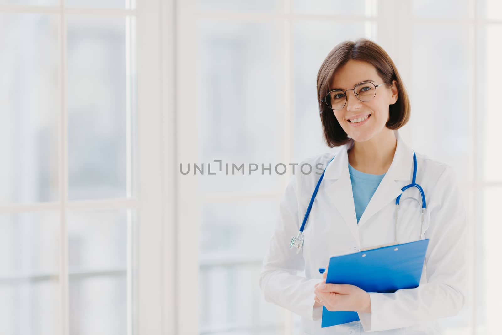 Good looking female expert creats medical prescription, writes down information in clipboard, thinks about good advice for patient, stands indoor, copy space for your information. Medicine concept by vkstock