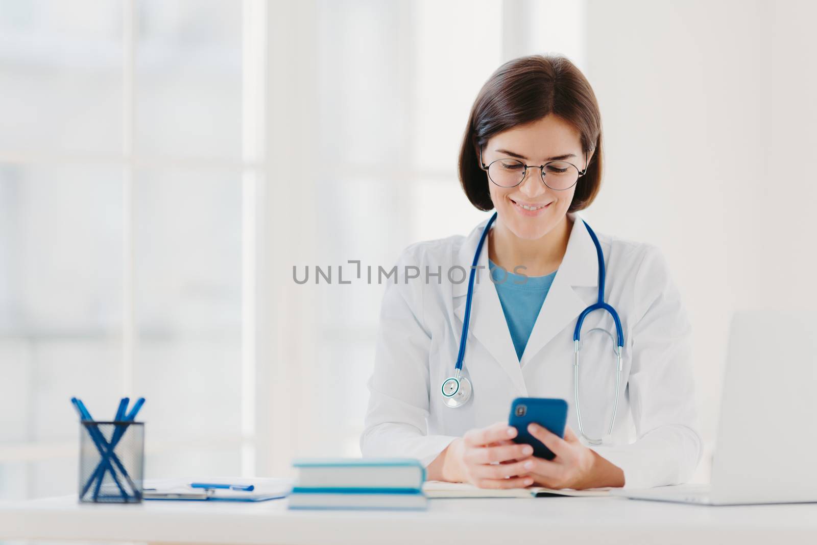 Female medical worker uses smartphone, reads advice how to cure disease in internet, poses in medical office, wears special medical uniform, sees results of examination for patient, examines chart