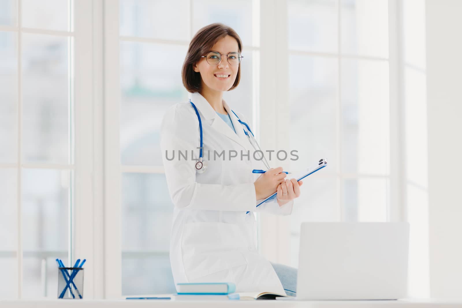 Indoor shot of professional female doctor supplies medic care assistance, poses at desktop with laptop, prepares document agreement paper offer, wears optical glasses and white medical gown. by vkstock