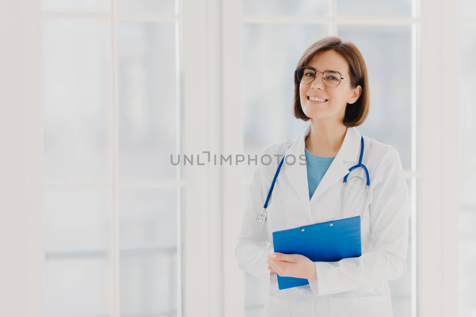 Experienced woman pediatrician stands with clipboard in cabinet, wears white medical coat with stethoscope, gives excellent medical treatment, has happy expression, ready to give consultancy