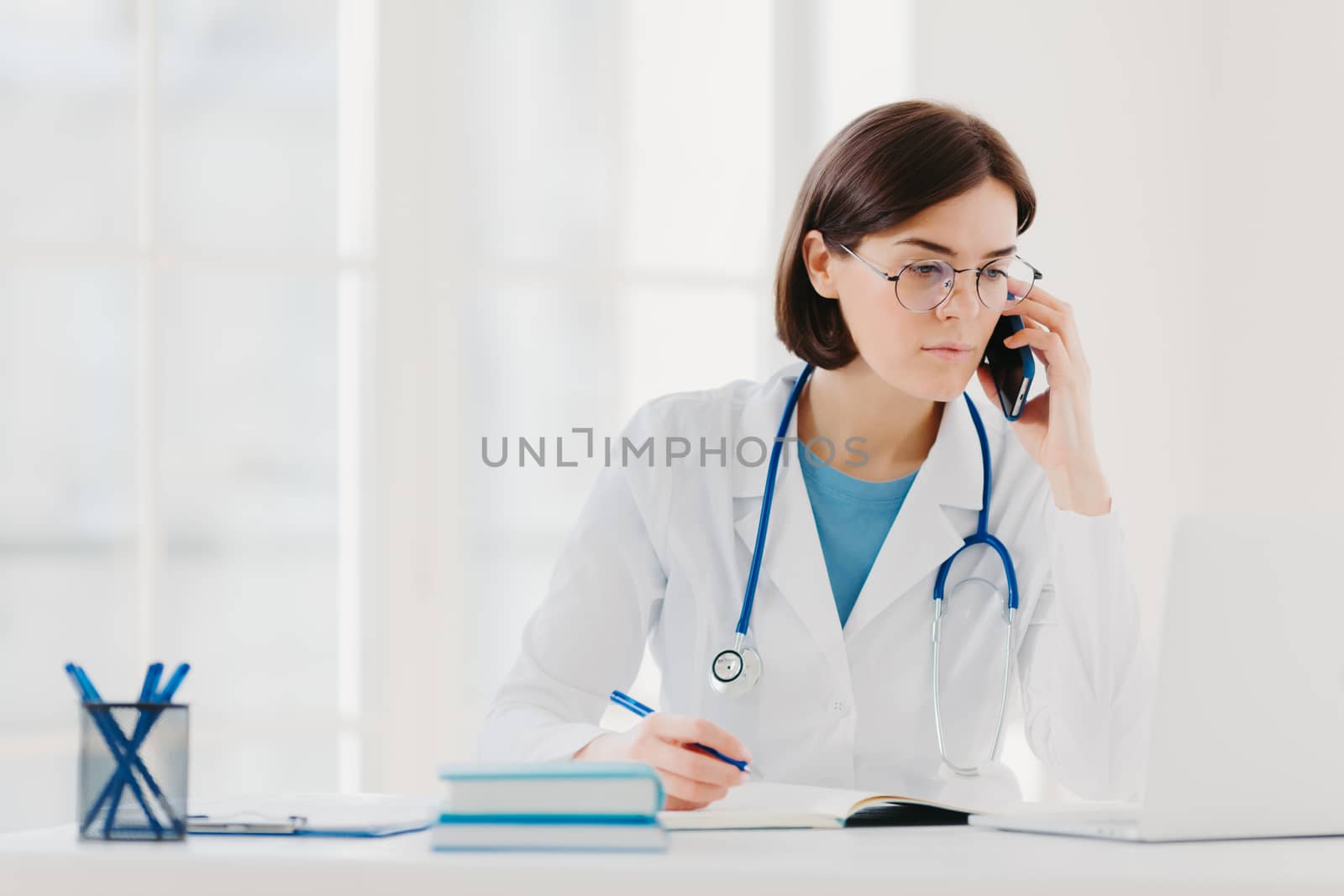Heathcare personnel, medicine concept. Serious brunette female doctor focused at modern laptop computer, rewrites necessary information, talks on mobile phone, calls someone, has serious look by vkstock