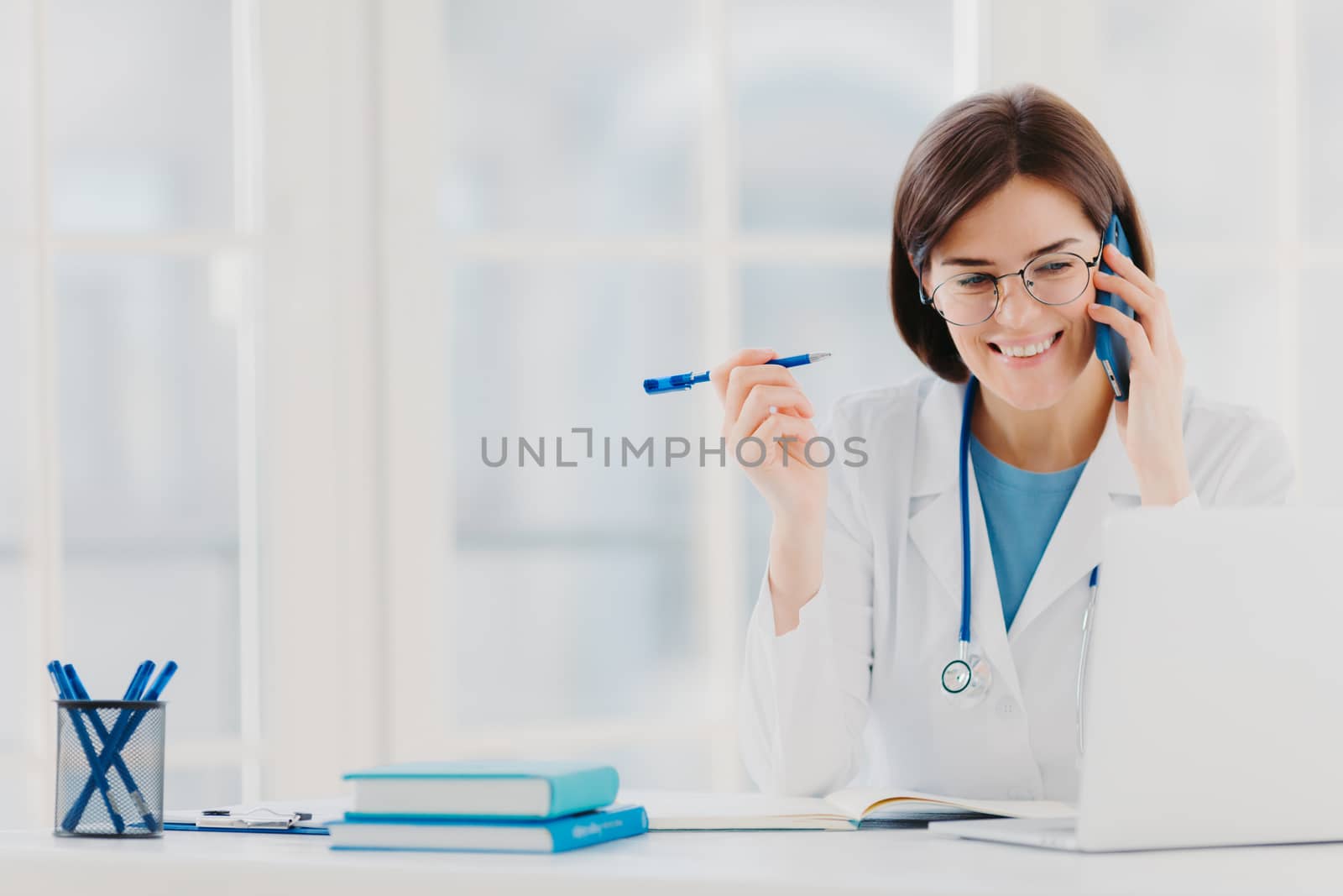 Glad professional doctor concentrated in modern laptop computer, reads useful information, has telephone conversation, discusses medical issues, sits at hospital office with notepads on table