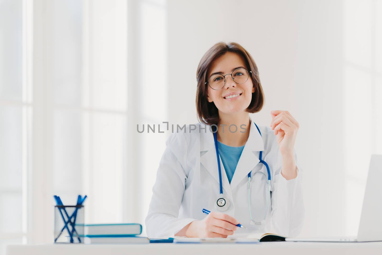 Young woman doctor writes on paper in clinic, creats prescription for patient, does paperwork in hospital, wears medical white gown, sits at cozy modern office. People, occupation, job concept