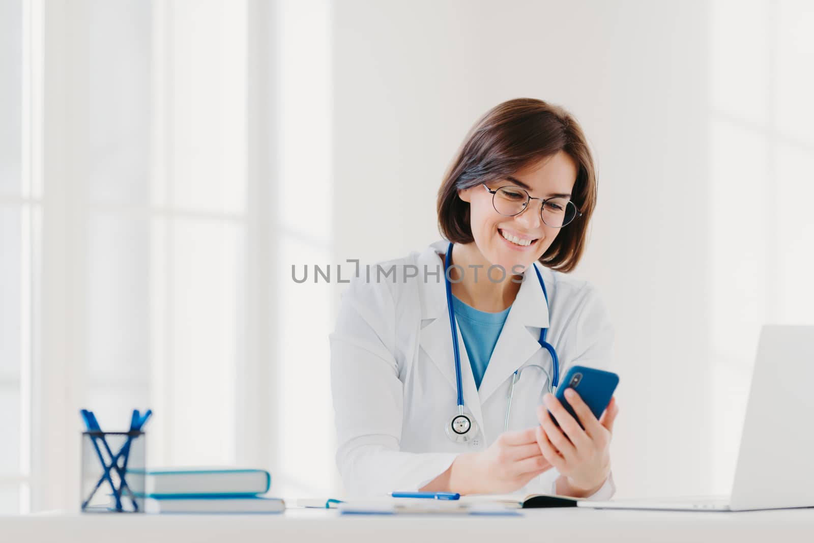 Positive female medical worker involved in chatting, holds mobile phone and sends messages, wears medical gown, has friendly cheerful expression, poses at desktop gives online consultation for patient