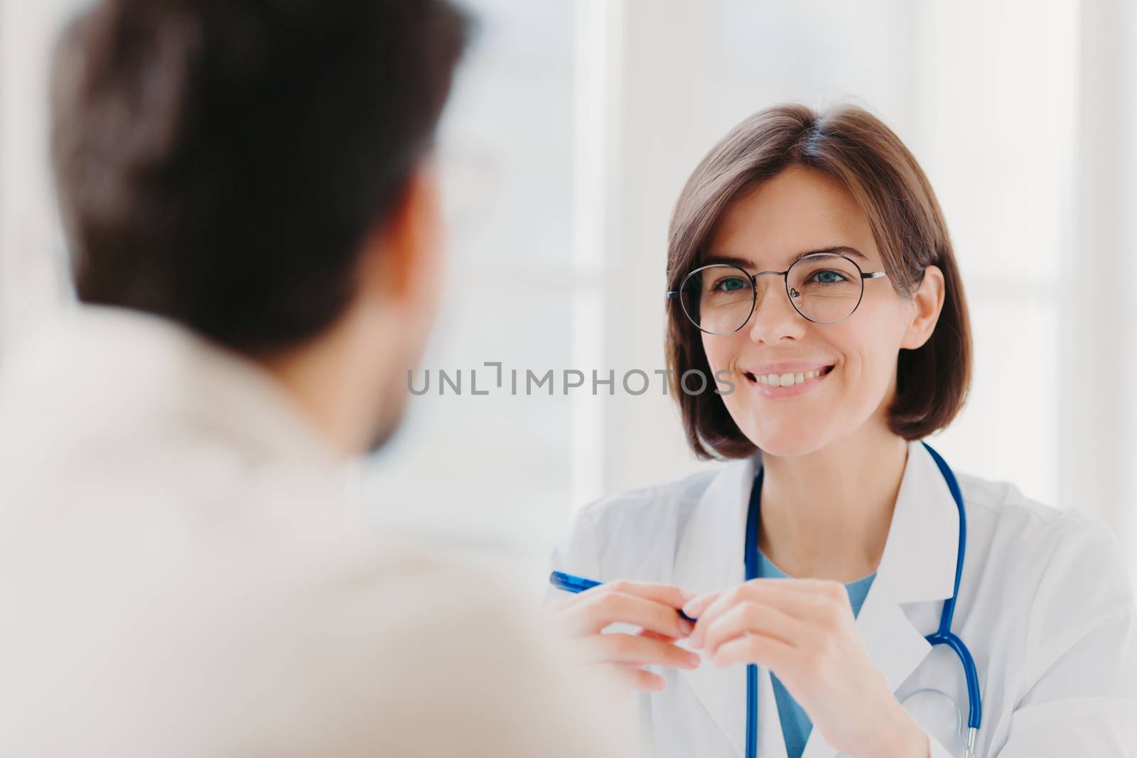 Smiling brunette woman doctor in round glasses, wears white coat, has conversation with patient, happy to help other people, wears white coat uniform, discuss health care. Good treatment concept by vkstock