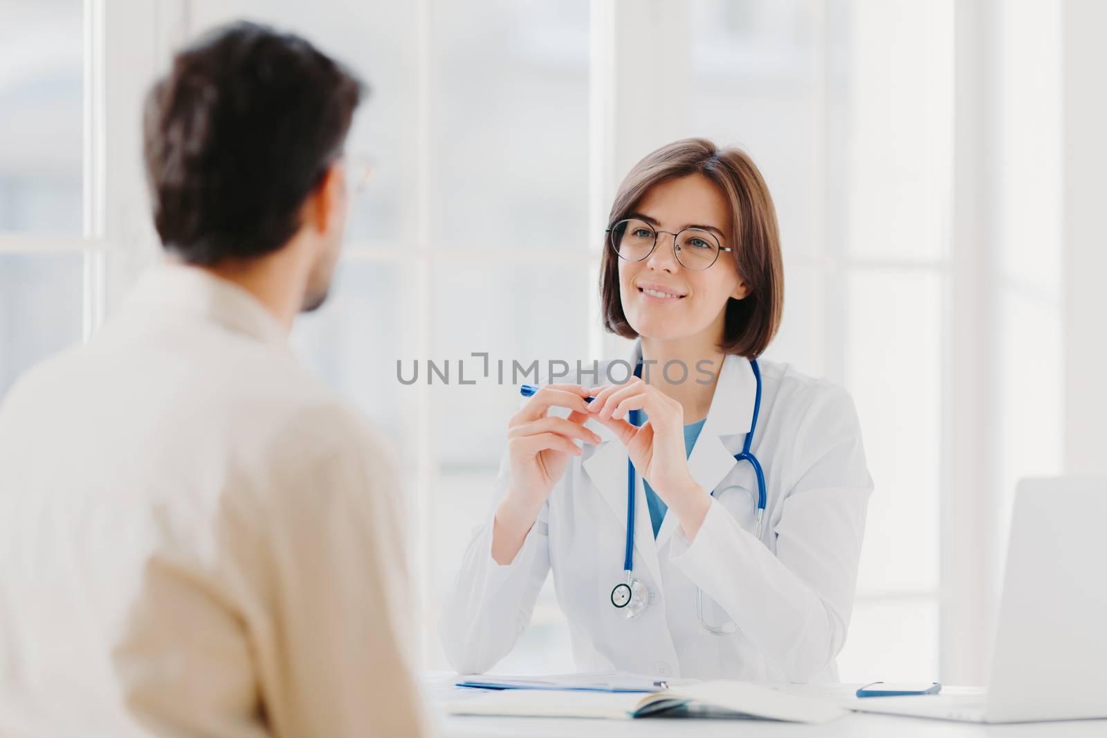 Professional female doctor in coat listens attentively patients symptoms, examines health, writes down information in patient card, pose in clinic. Sick male tells complaint, sits back to camera by vkstock