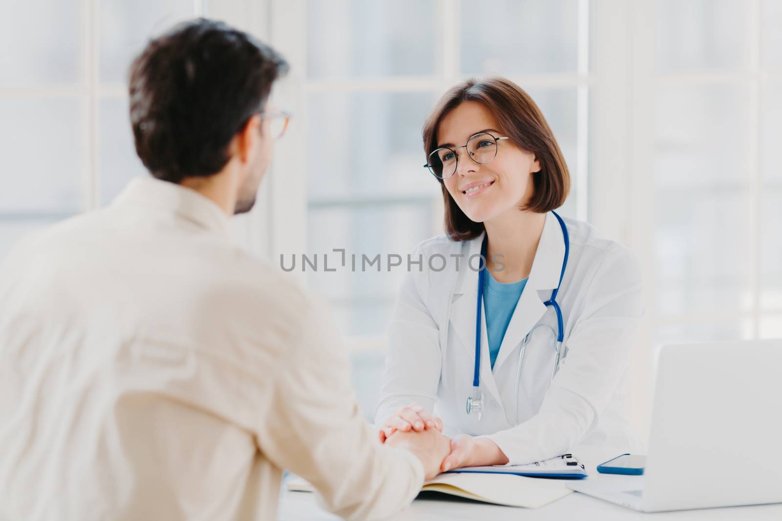 Confident female doctor holds hands of ill patient, persuades everything will be alright, dressed in white medical gown, gives advice, pose in hospial office. Consultation and diagnosis concept