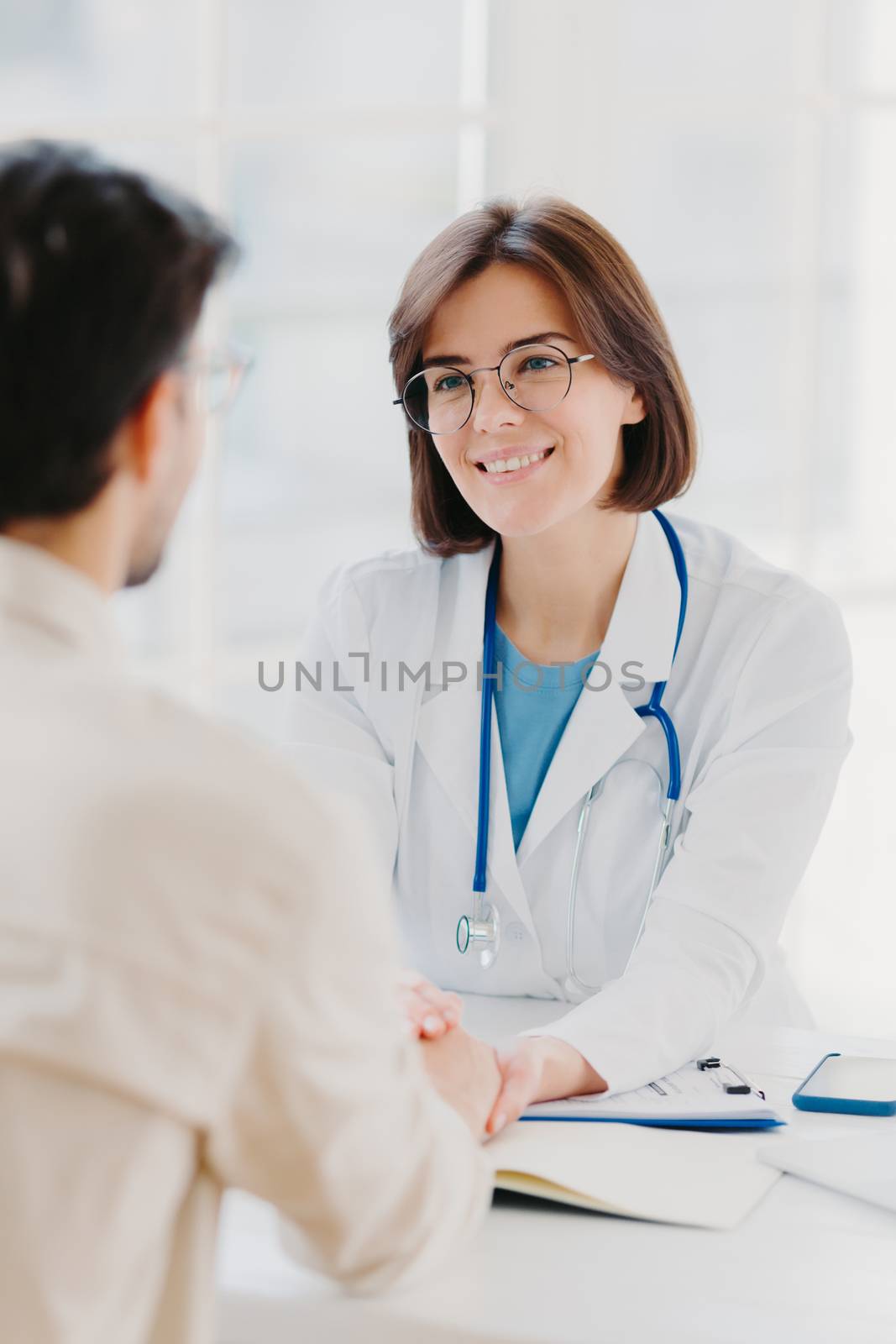 Medical trust concept. Vertical image of female doctor reassures her male patient, hold hand and talk calmly about illness cure, gives professional consultation and support, pose at doctors office by vkstock