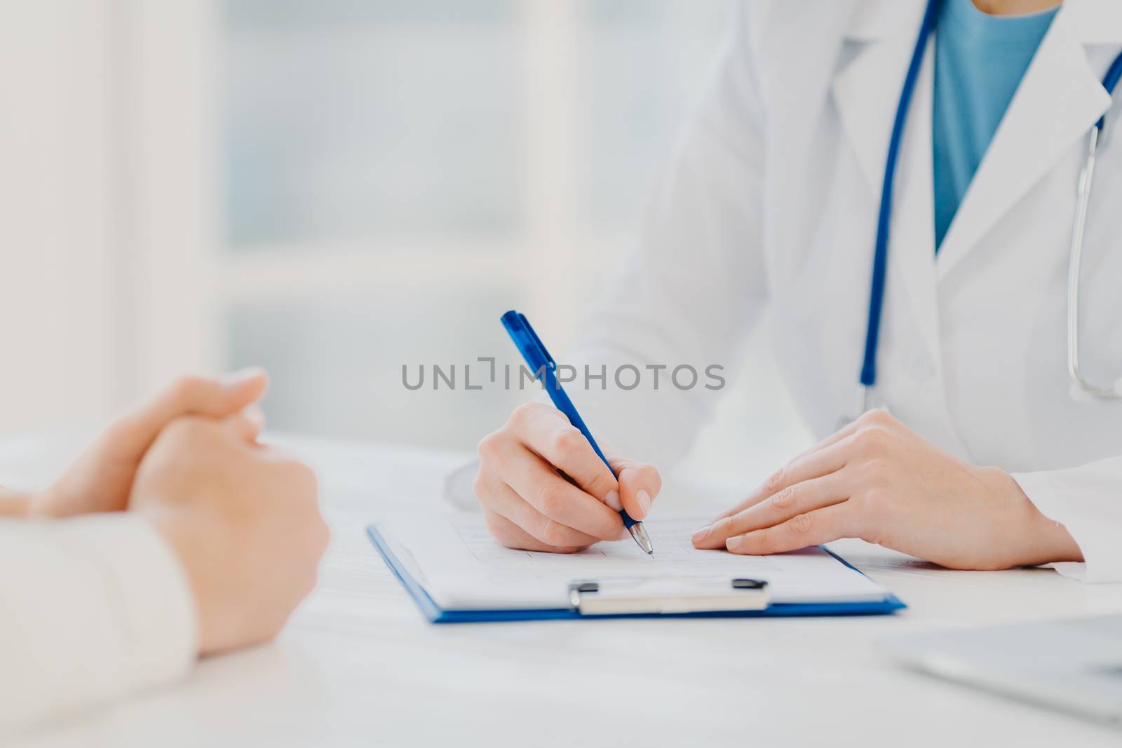 Unknown female doctor writes down prescription on papers in clipboard, consults patient about curing disease, pose at desktop against blurred white background fills up medical form. Symptoms diagnosis