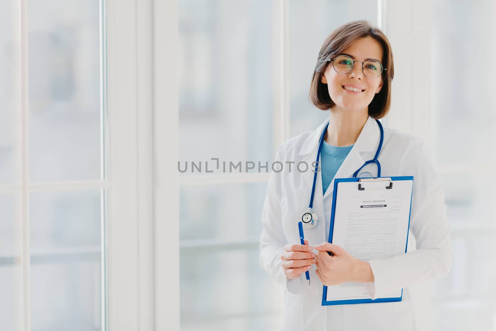 Horizontal shot of woman doctor stands with clipboard, fills up application form, holds pen, smiles positively, enjoys her work, helps people, stands in white medical gown against big window by vkstock