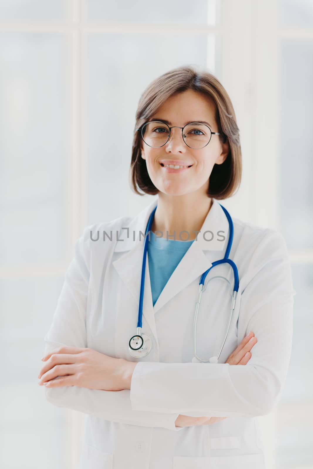 Beautiful cheerful woman doctor wears white gown, glasses and phonendoscope, keeps hands crossed, looks confidently at camera, has short dark hair, stands against white background, works in clinic