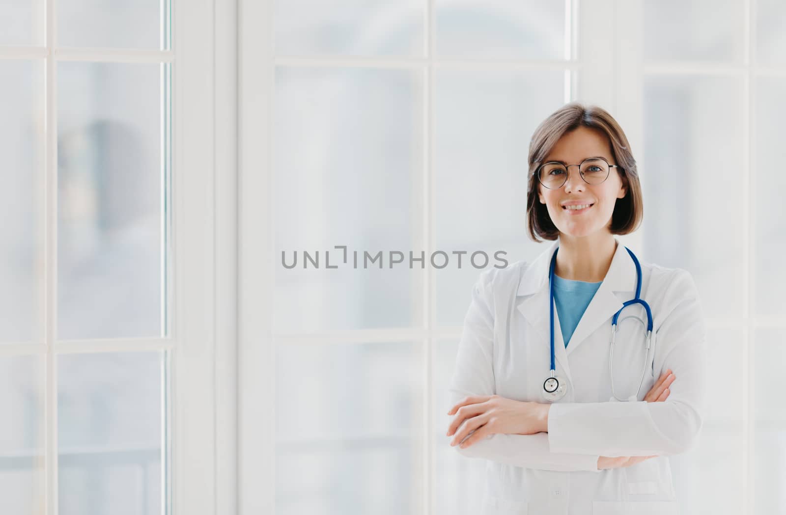 Self confident professional doctor stands with arms crossed, wears white medical gown with stethoscope, thinks about work positively, poses against big window. Healthcare and occupation concept by vkstock