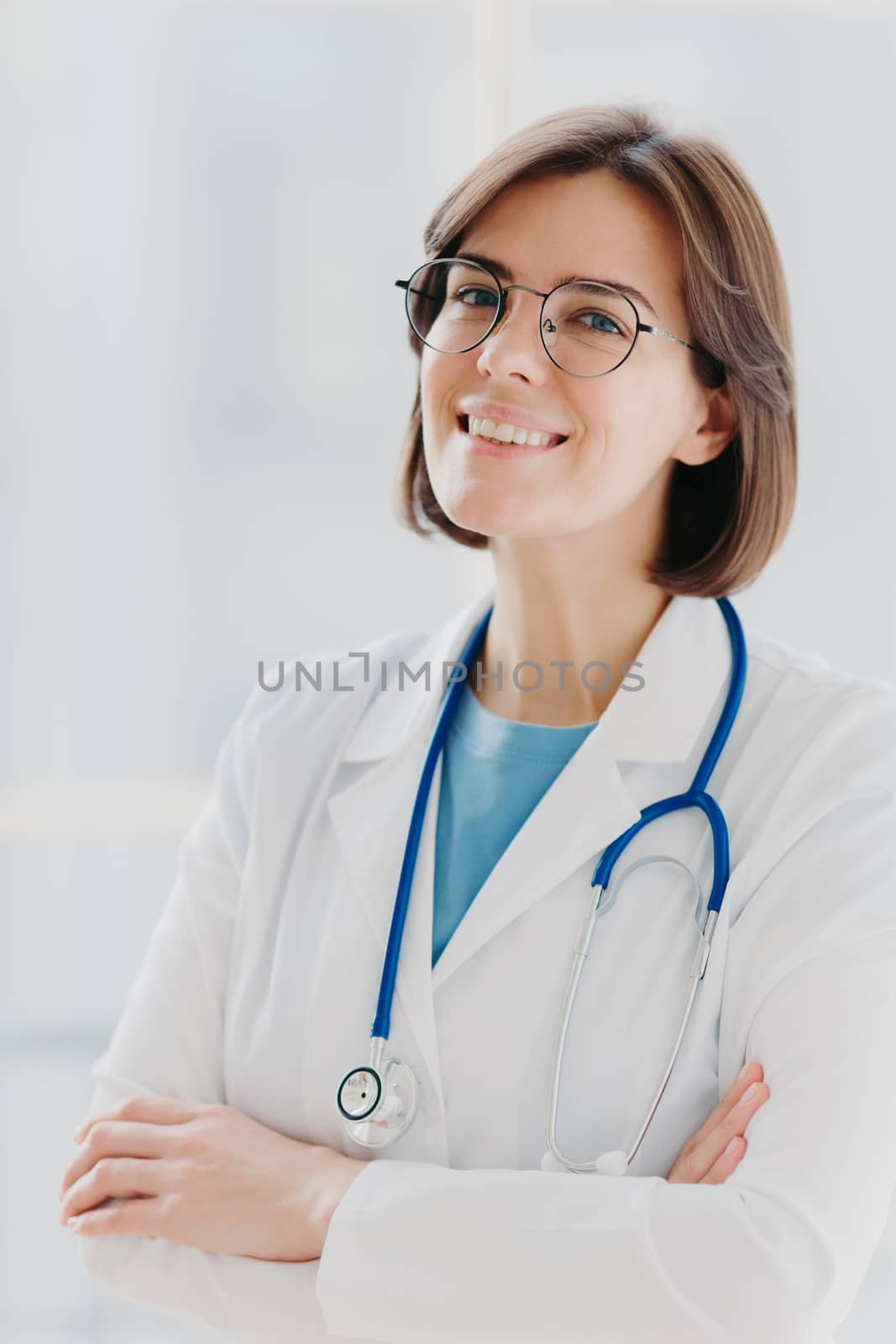 Close up portrait of short haired female general practitioner stands with smile and arms crossed, uses stethoscope, enjoys work, poses against white background. People, mediccare and treatment concept by vkstock