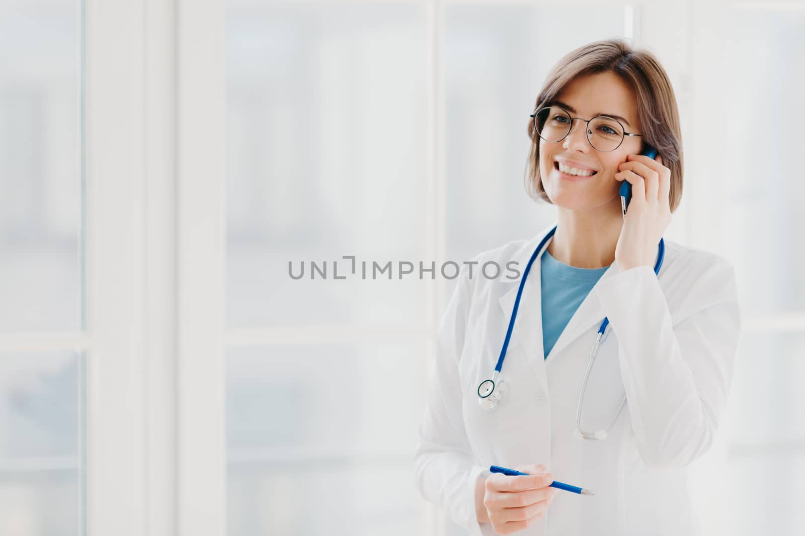 Shot of pleased female physician or surgeon gives prescription via mobile phone, has conversation with patient, holds pen, wears white uniform, works in hospital, stands indoor against window by vkstock