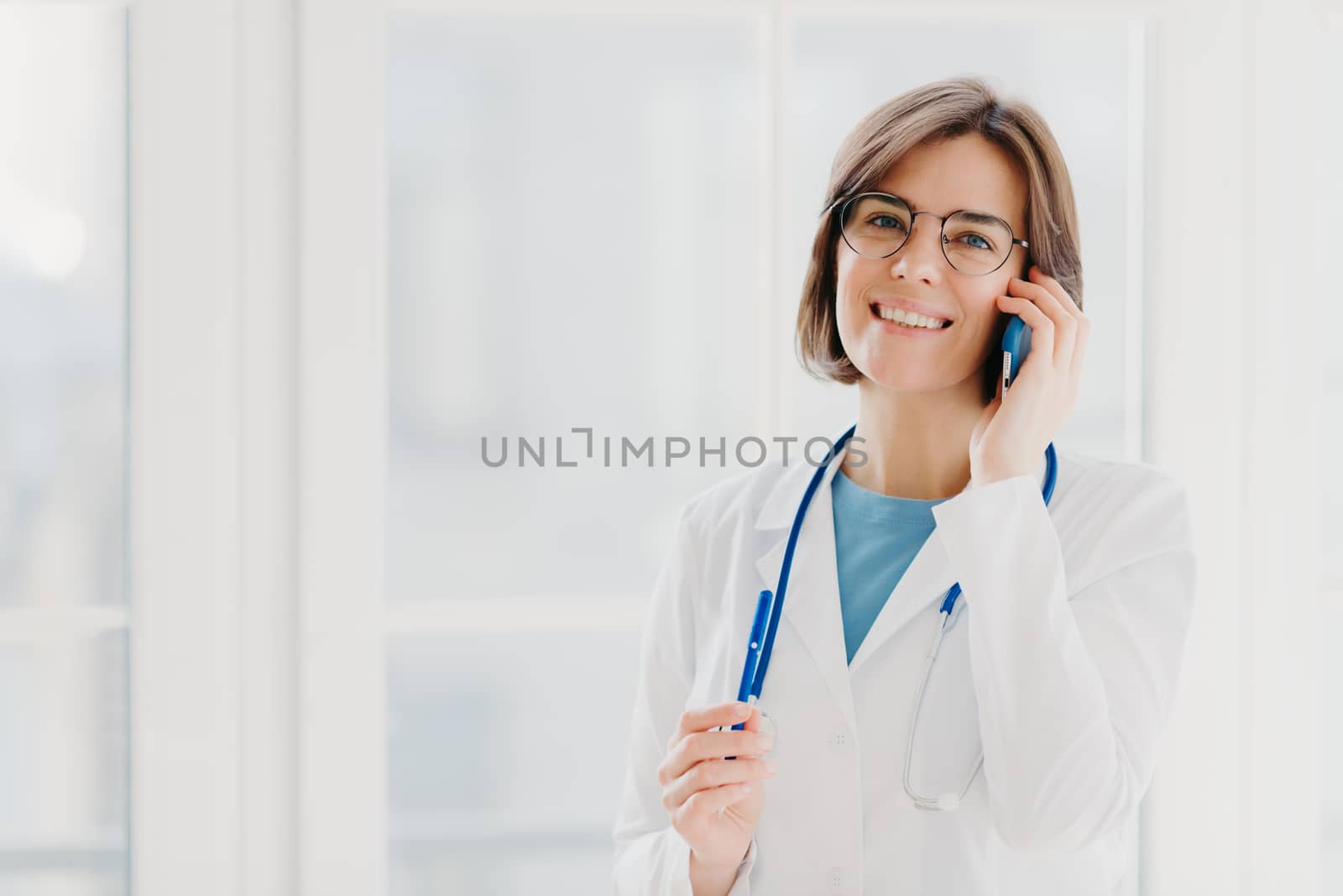 Positive woman doctor consults patient via cellphone, looks gladfully at camera, holds pen, wears white coat, poses in medical office, poses near window. Healthcare and consultation concept. by vkstock