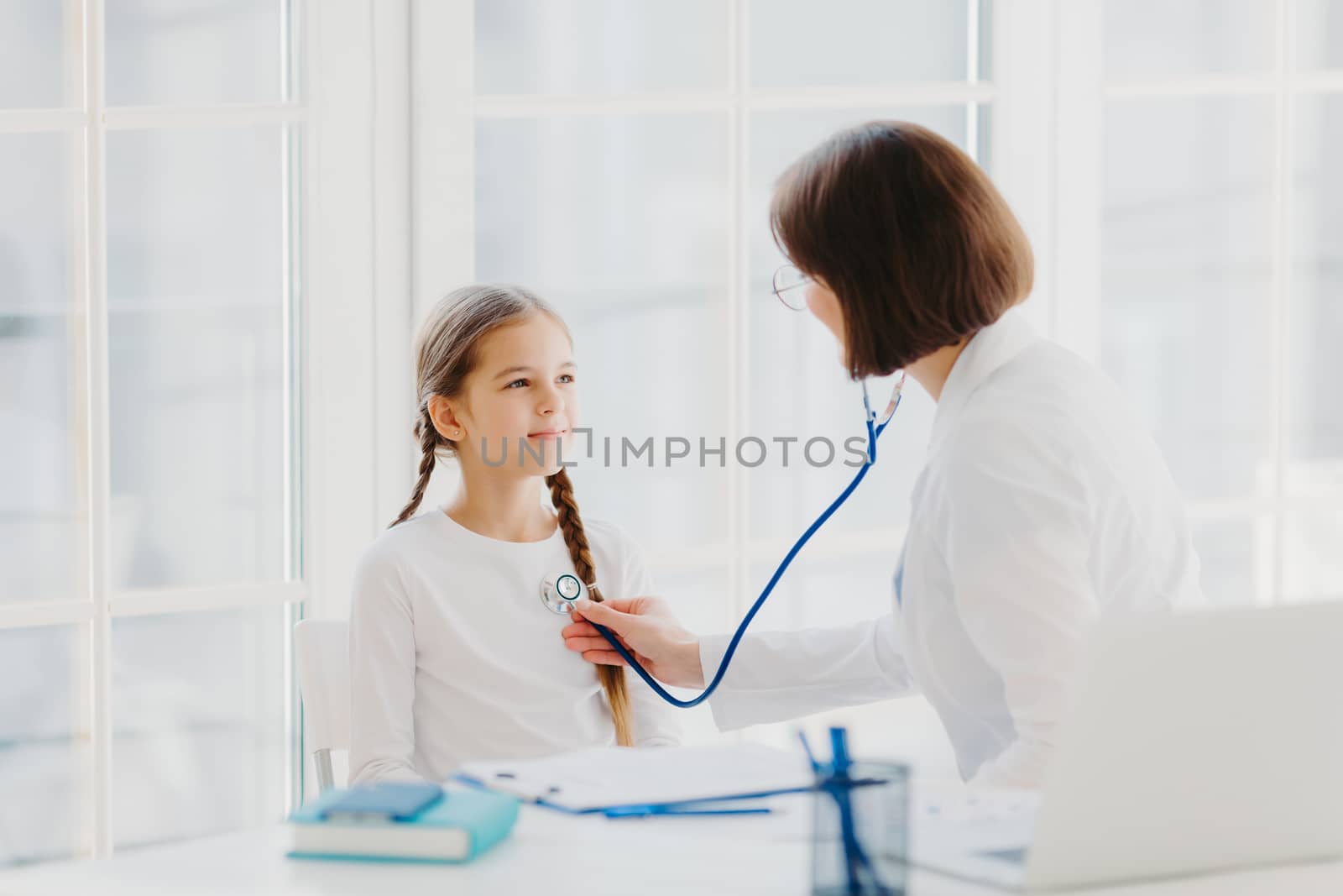 Woman pediatrician gives consultation to small girl, talk about health and symptoms, listens heart with phonendoscope, makes prescription, pose in hospital office. Childrean medical insurance and care by vkstock