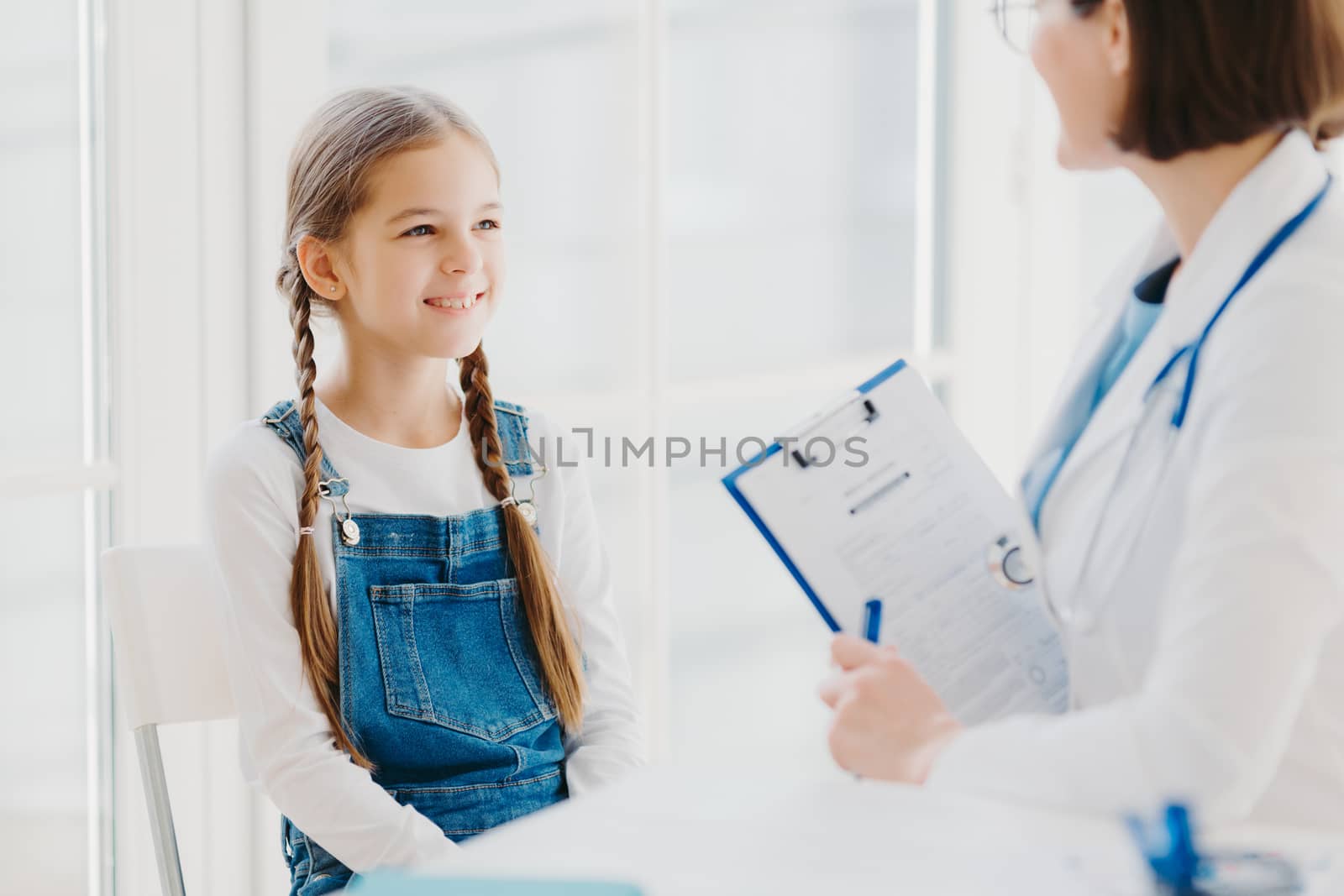 Happy little child listens attentively doctors advice and prescription, comes to see pediatrician in clinic, has two pigtails, wears denim overalls, talk about health problems, has checkup examination by vkstock