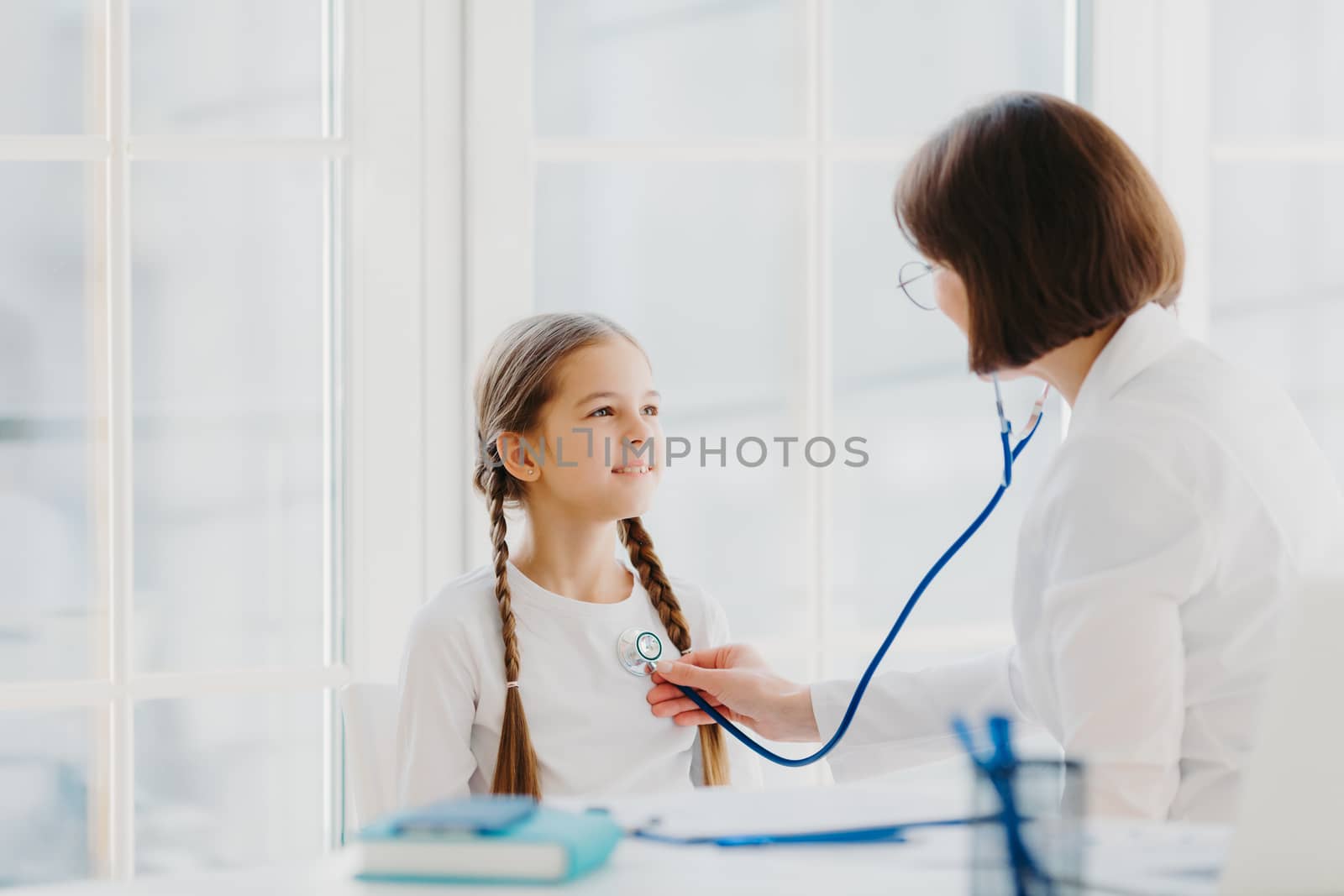 Lovely small girl listens consult of professional experienced doctor who listens her lungs with stethoscope, comes on medical checkup appointment. Children healthcare and clinic visit concept by vkstock