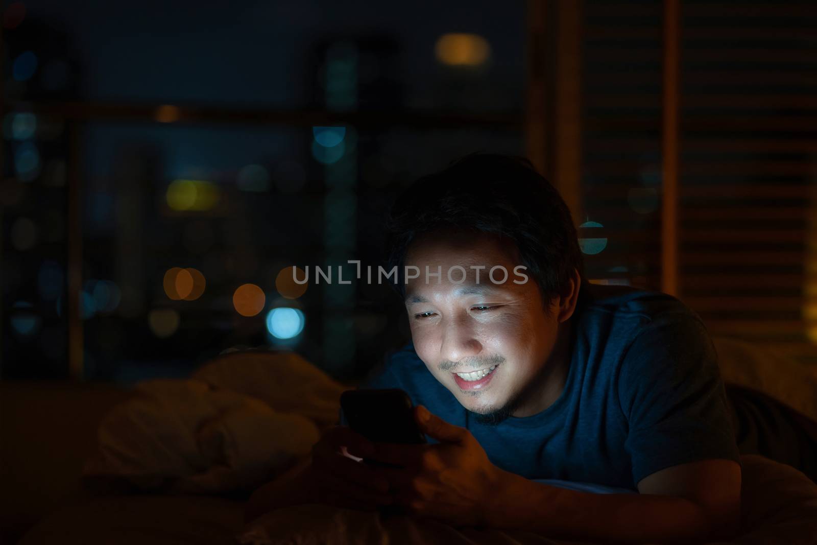 Closeup Asian man sleeping and using smart mobile phone for social network or video conference call and face time to her lovers on bed over blur city at night time, lifestyle and relax, work from home