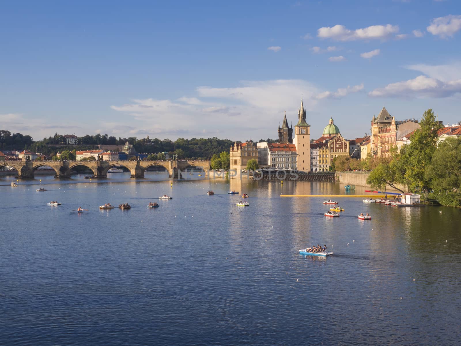 panorama of Charles bridge over Vltava river and houses of Old Town, Prague, Czech Republic, golden hour light, summer sunny day, tourists relaxing on pedal boats.
