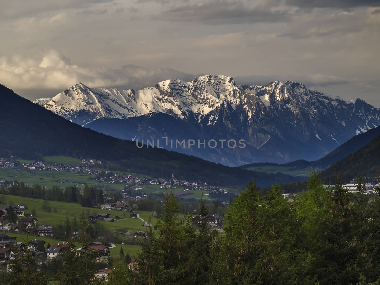 Spring mountain rural landscape. View over Stubaital Stubai Valley near Innsbruck, Austria with village Neder, green meadow, forest, snow covered alpen mountain peaks, evening dramatic clouds and light.