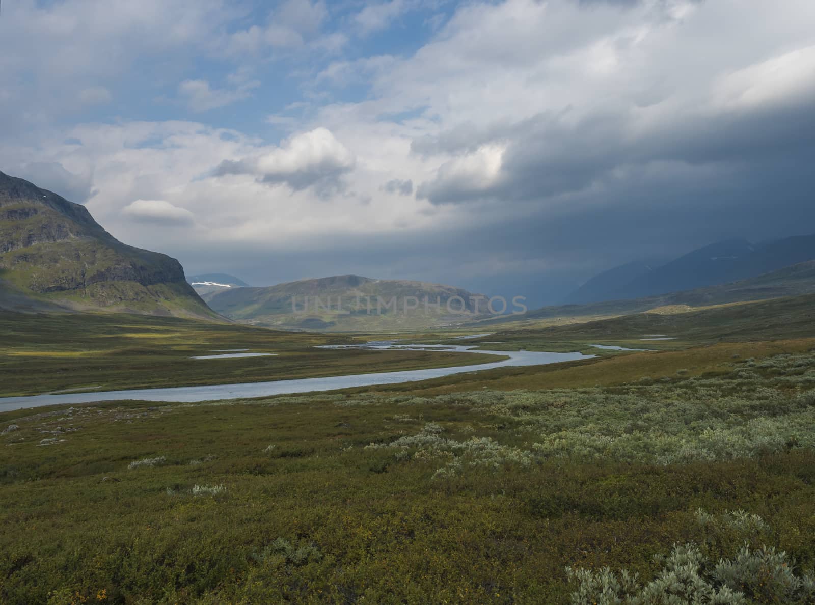 Beautiful wild Lapland nature landscape with blue glacial river, birch tree bushes, snow capped mountains and dramatic clouds. Northern Sweden summer at Kungsleden hiking trail