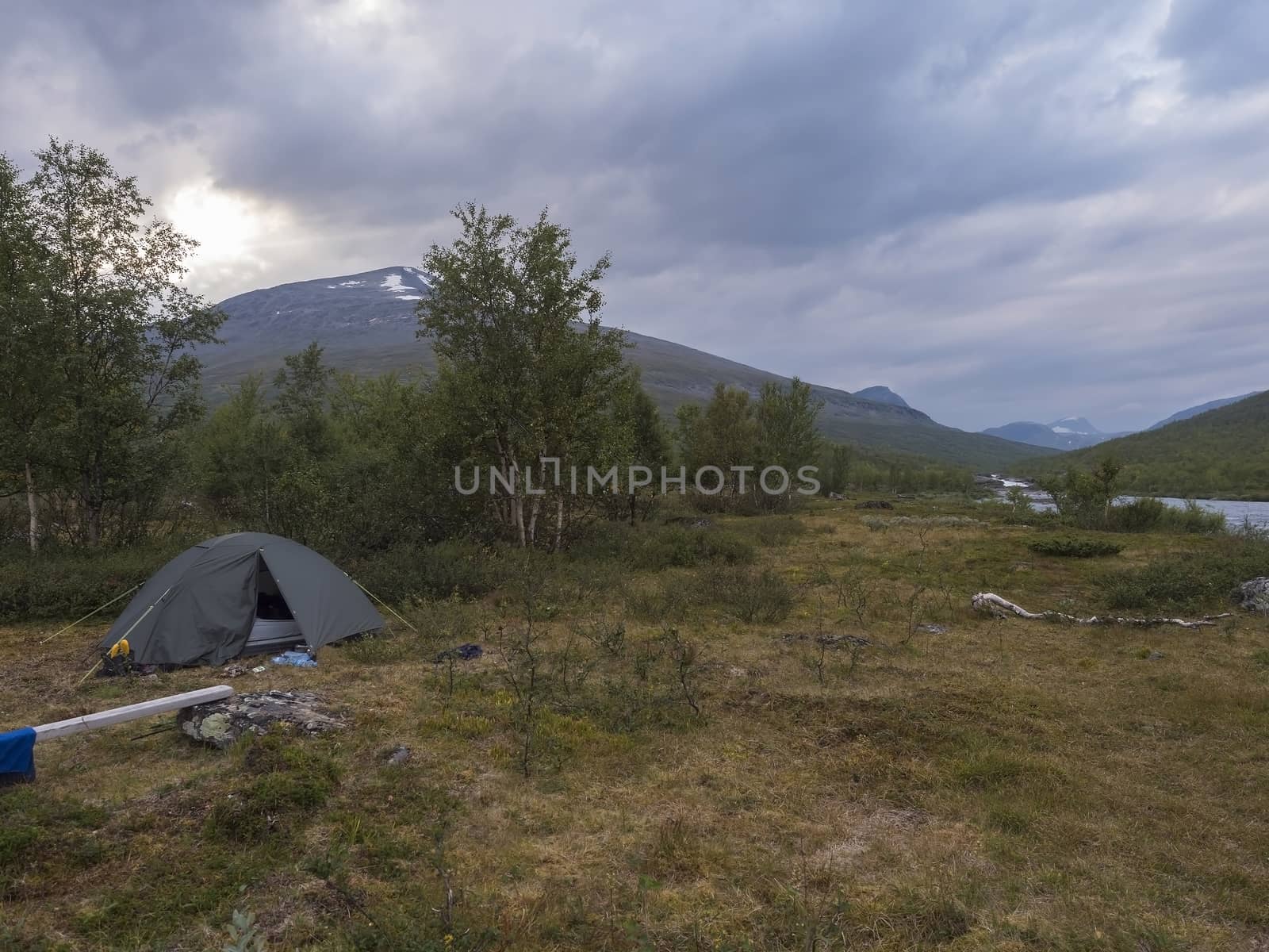 Small green tent in Beautiful wild Lapland nature landscape with blue river, Kaitumjaure lake, birch trees and mountains. Northern Sweden summer at Kungsleden hiking trail. Blue sky dramatic clouds by Henkeova