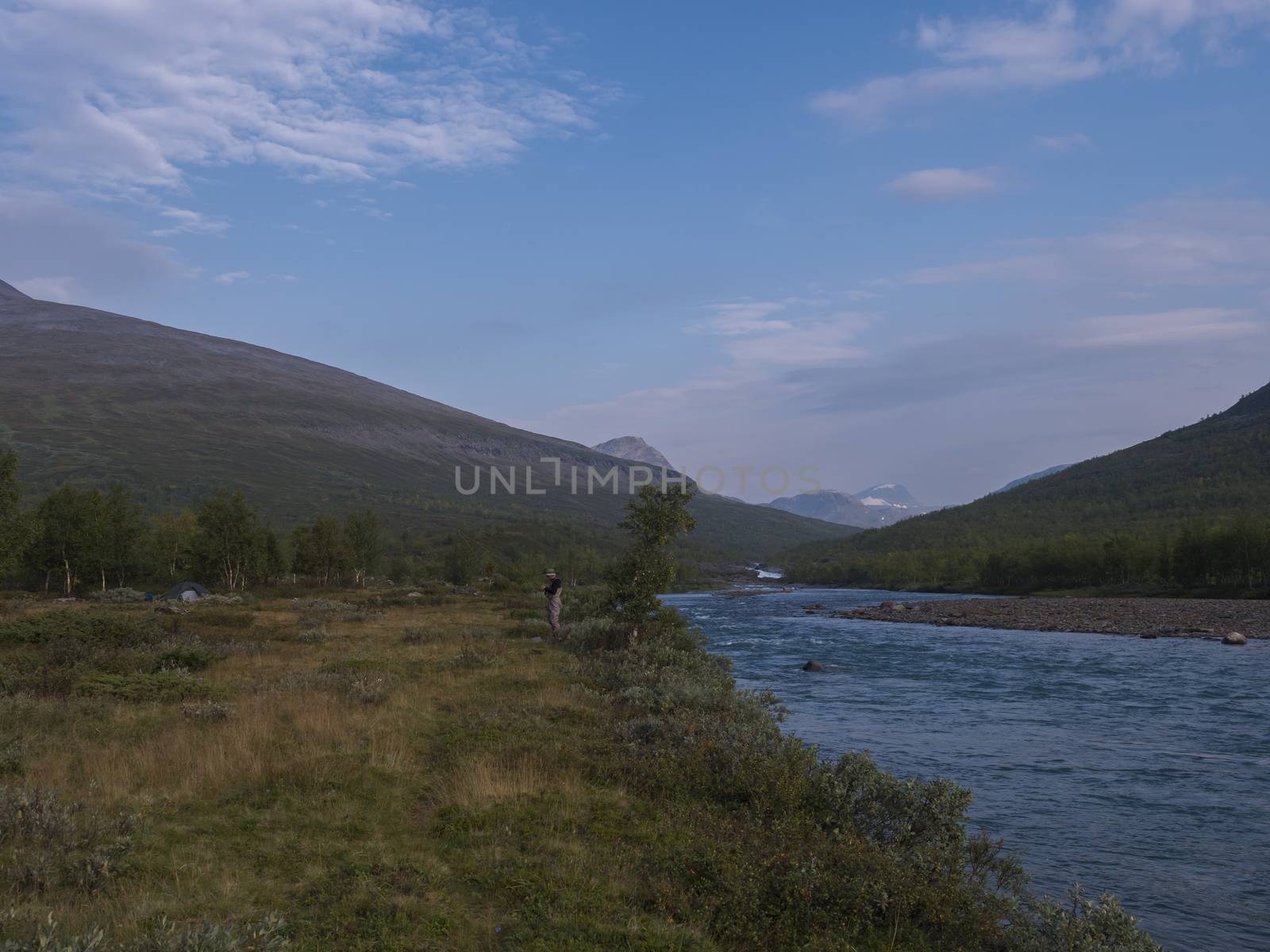 Wild Lapland nature landscape with angler fisherman at blue Tjaktjajakka river, Kaitumjaure, birch tree forest and mountain Sanjartjakka. Northern Sweden summer at Kungsleden hiking trail. Blue sky background.
