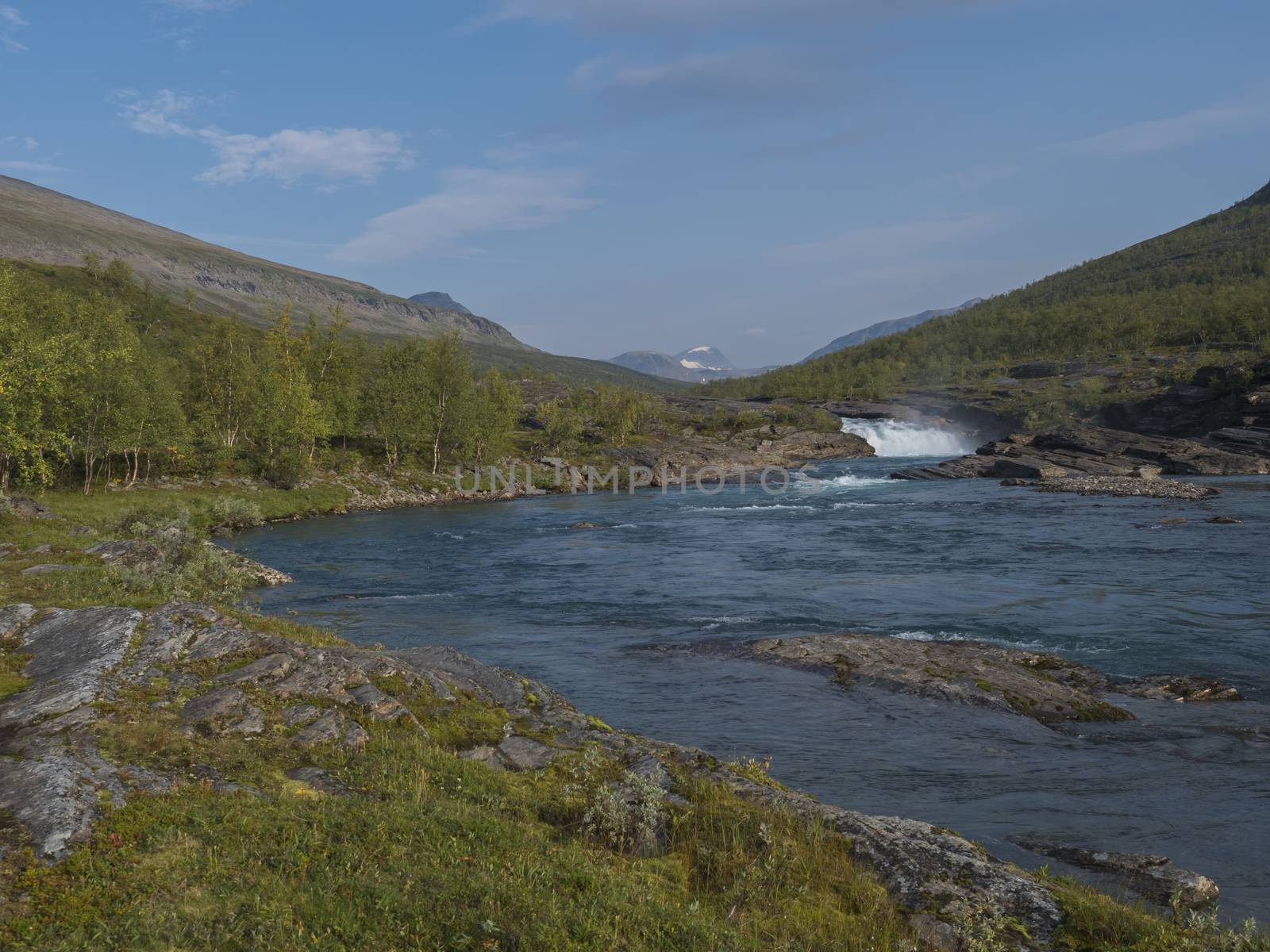 Beautiful landscape with waterfall on wild Tjaktjajakka river, birch tree forest and mountains. Lapland naturein summer at Kungsleden hiking trail. Blue sky background.