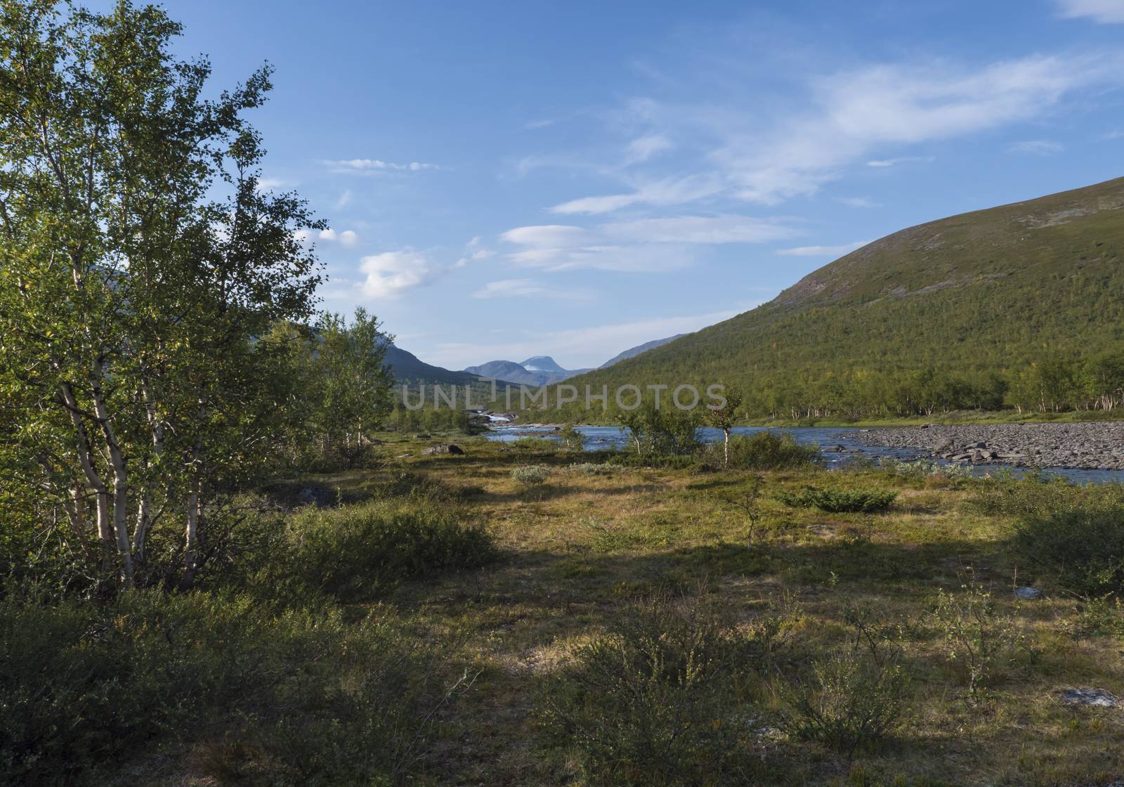 Beautiful landscape with waterfall on wild Tjaktjajakka river, birch tree forest and mountains. Lapland naturein summer at Kungsleden hiking trail. Blue sky background.