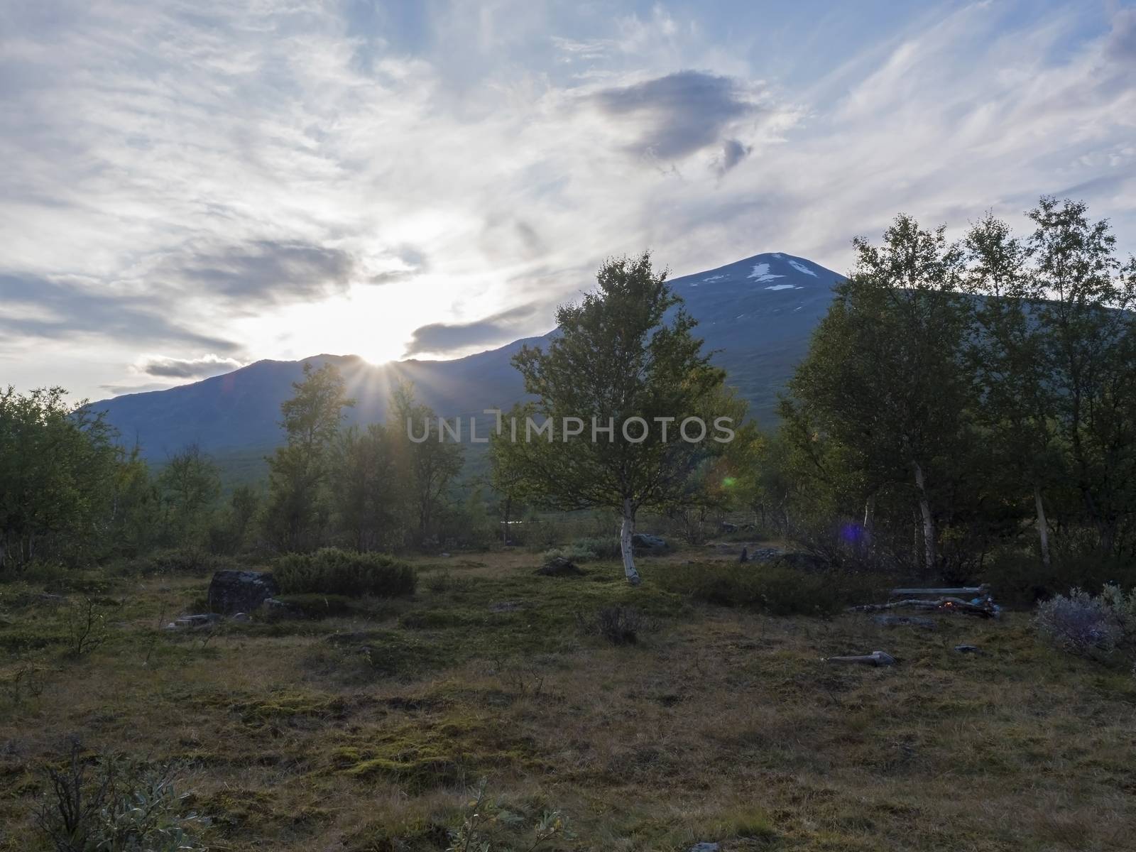 Sunset view on mountain Sanjartjakka with small camp fire and Beautiful wild Lapland nature landscape with birch tree forest. Backlight with lens flare.